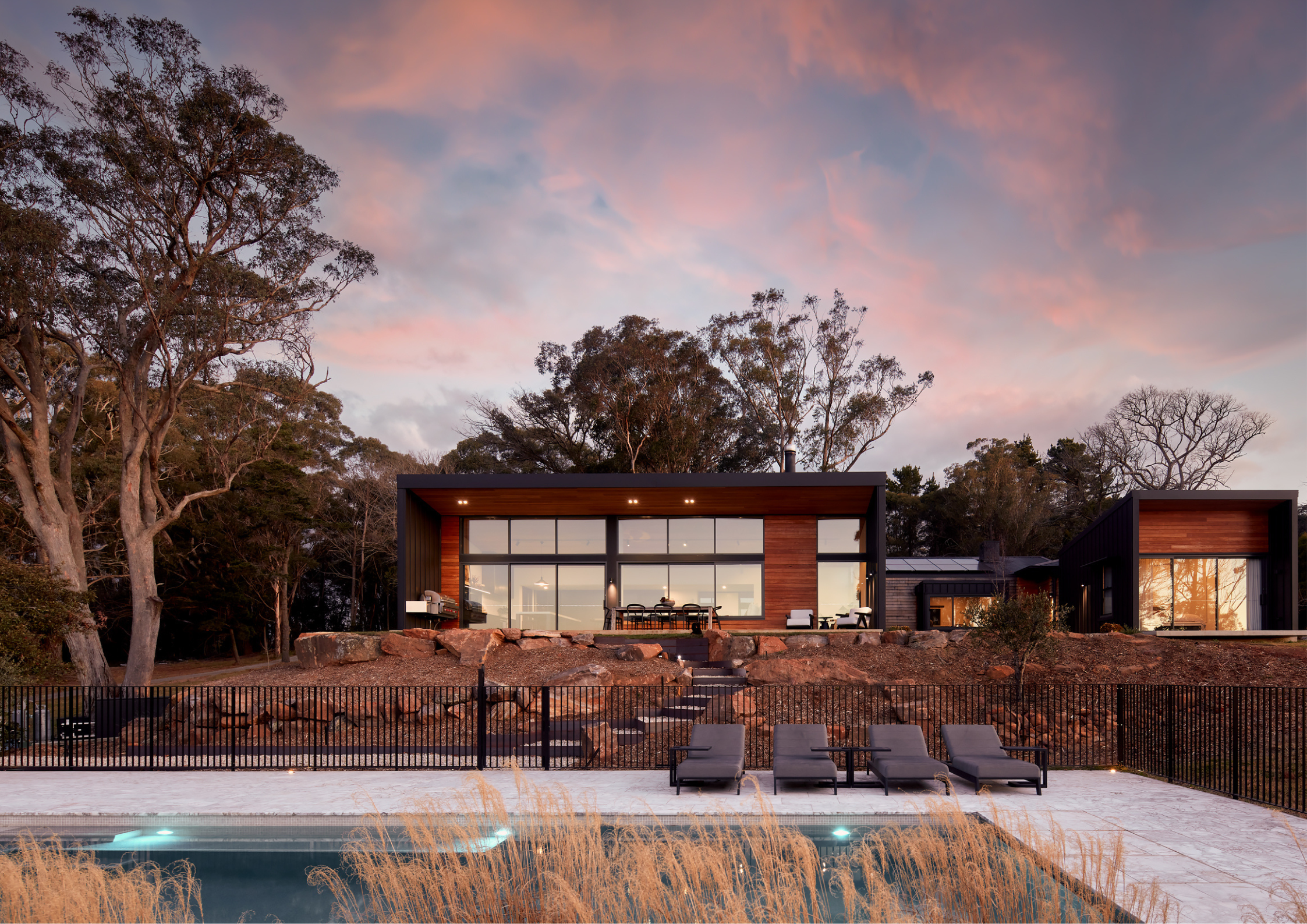 Contemporary pavilion-style home with expansive glazing, warm timber cladding, and a poolside terrace set against a dramatic sunset and bushland backdrop.