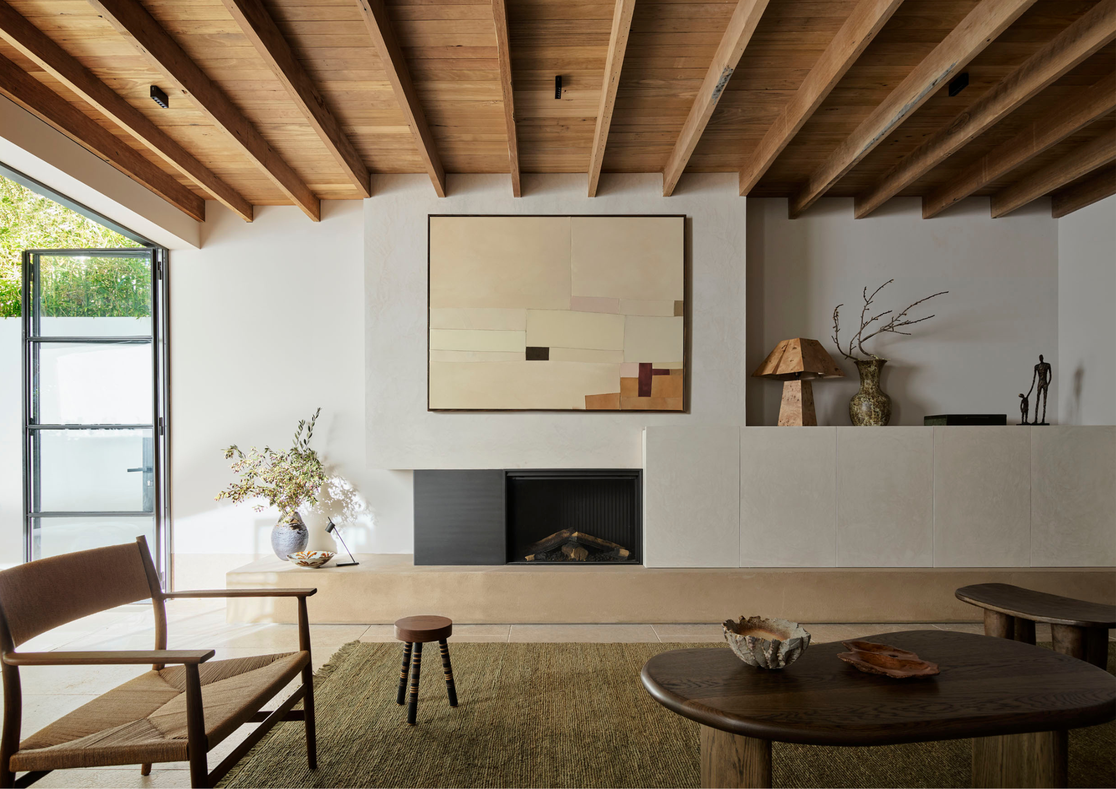 Minimal living room with exposed timber ceiling beams, sculptural furniture, a built-in fireplace and textured natural materials bathed in warm sunlight.
