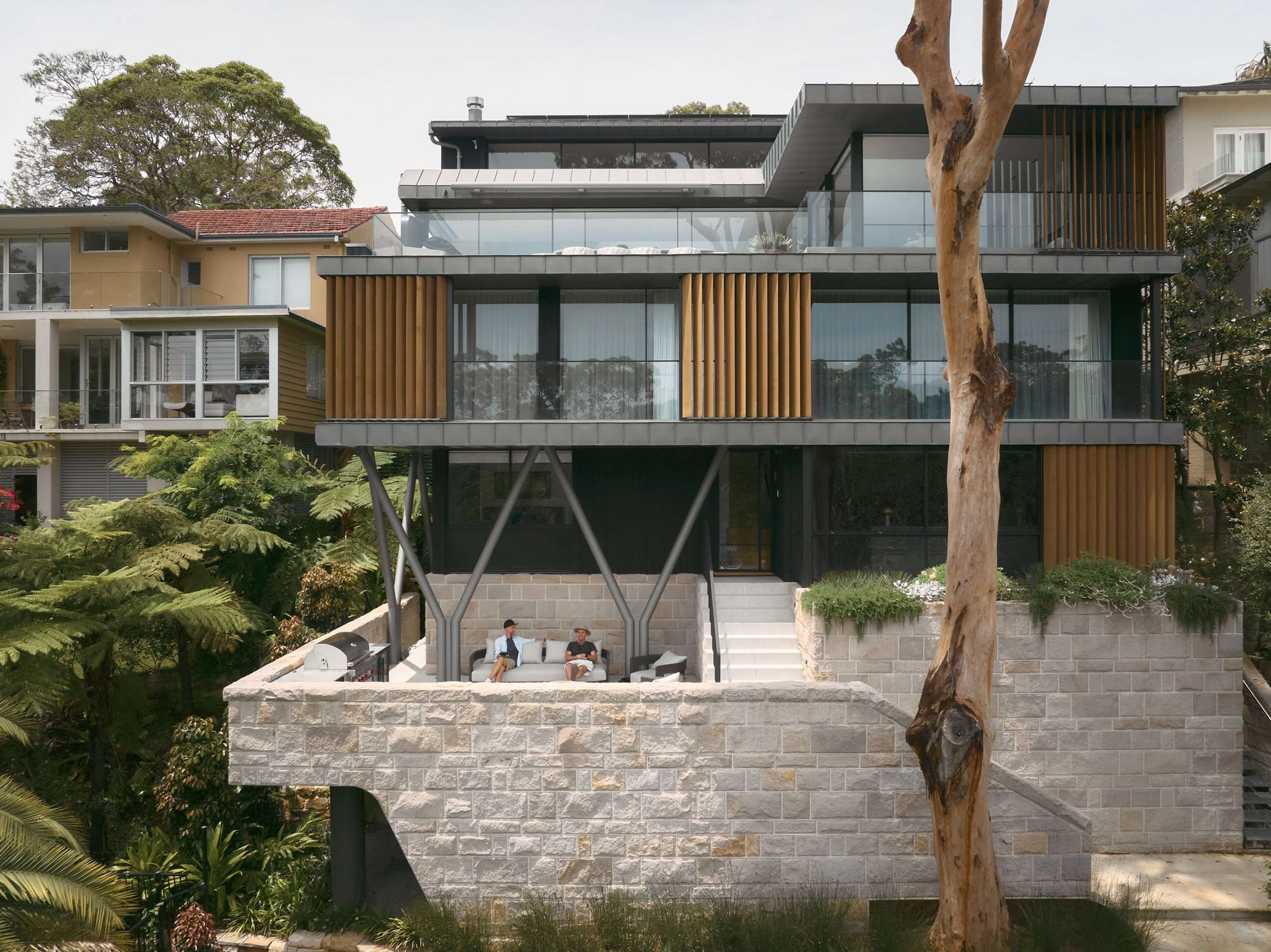 Multi-level contemporary waterfront home with stone retaining walls, vertical timber battens, and large glazed balconies overlooking lush greenery.