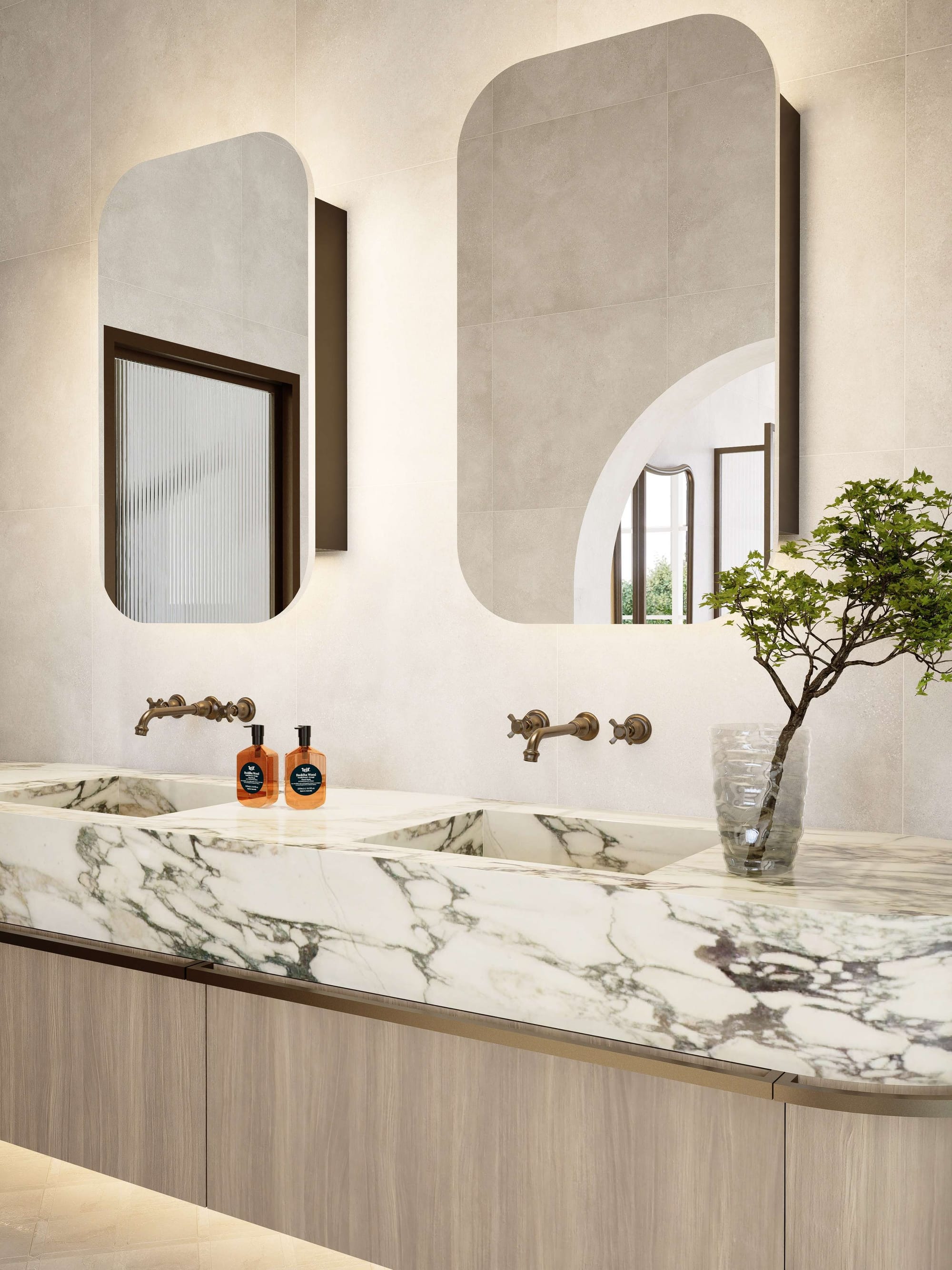 Contemporary bathroom vanity with curved marble double sink, brushed brass wall-mounted tapware, and twin rounded mirrors against soft stone-look tiled walls. Light timber cabinetry and a sculptural glass vase with greenery add warmth to the minimalist interior.