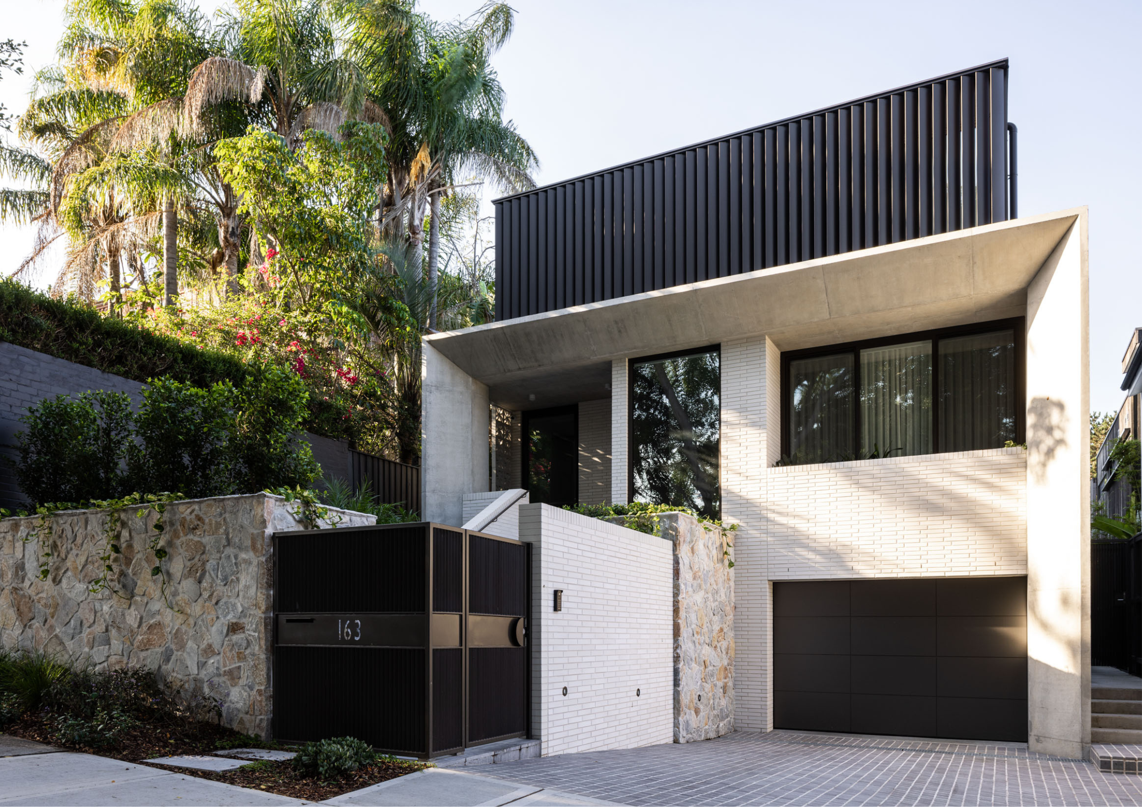 Angular contemporary home with white brickwork, exposed concrete, and dark vertical cladding set into a lush sloping landscape.