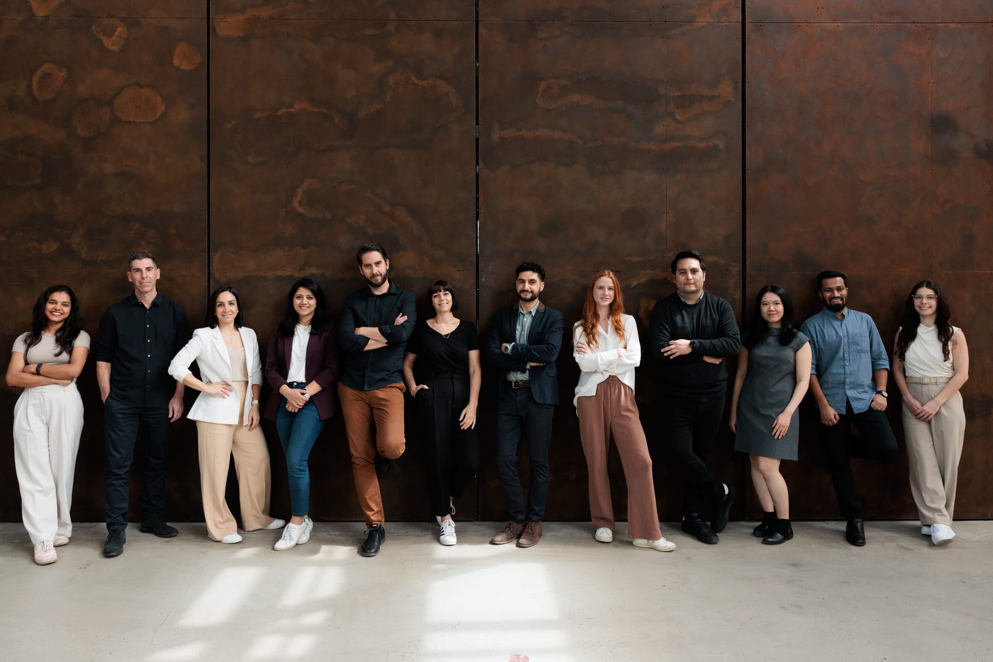 BJB Architects team standing together against a large rusted steel wall, dressed in smart casual attire in a warm, industrial setting.