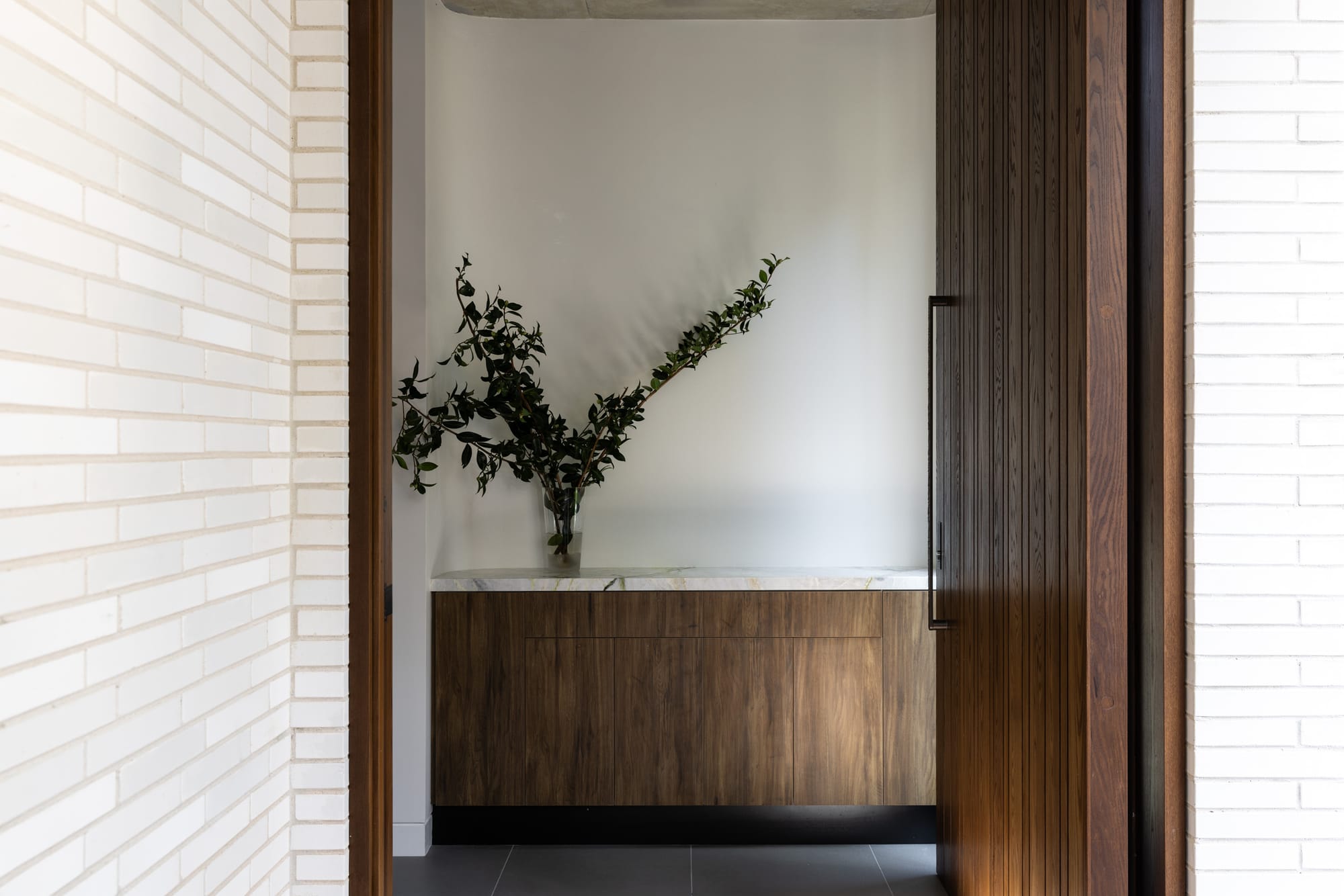 Minimal entry foyer with white brick walls, warm timber joinery, marble detailing and a sculptural branch arrangement beside a tall timber door.