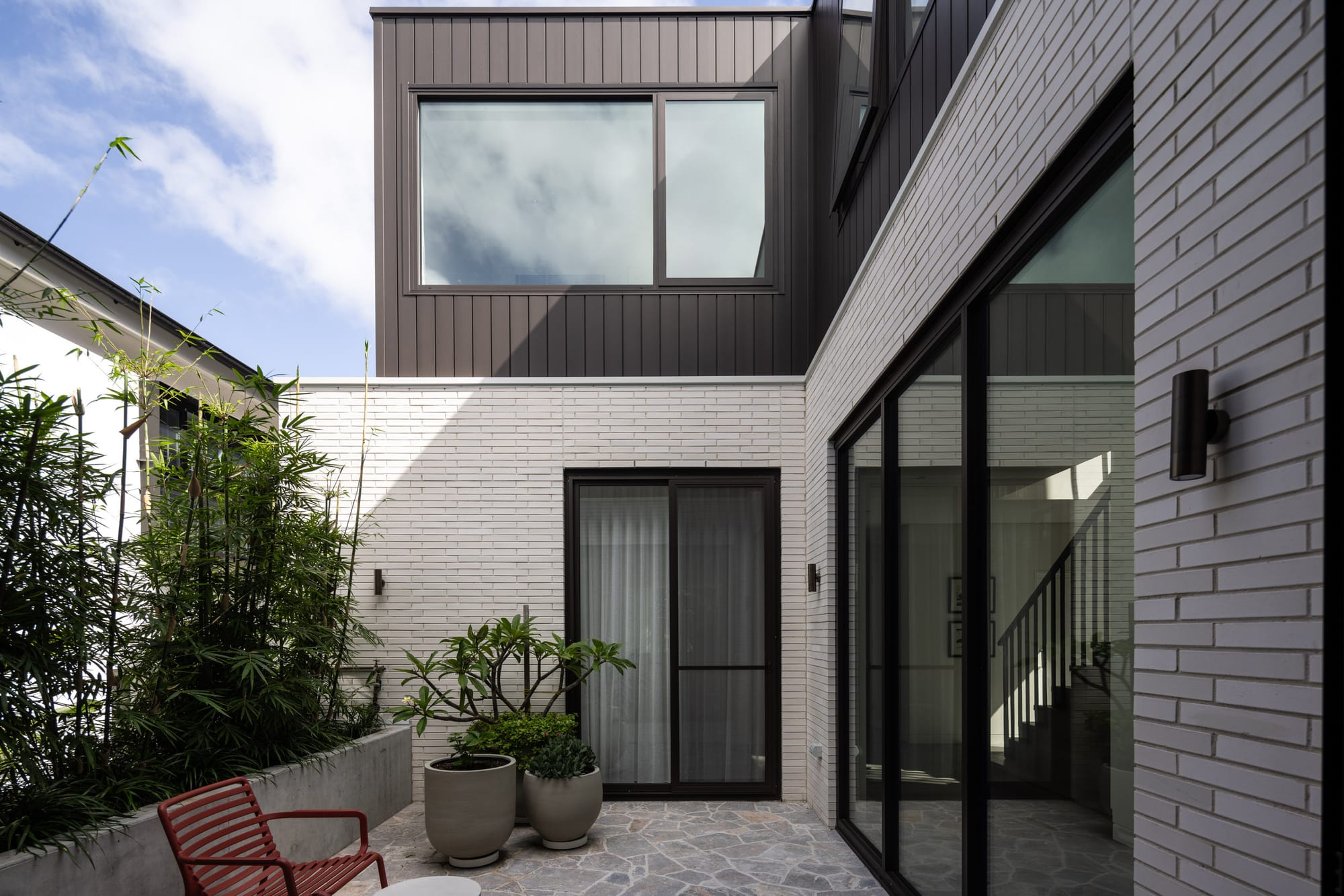 Compact courtyard framed by white brick and dark cladding, featuring large glazed openings, stone paving and lush greenery in oversized planters.