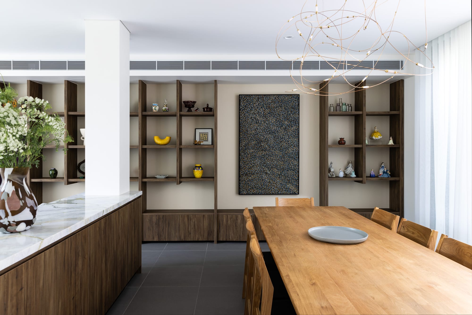 Refined dining space with warm timber shelving and furniture, marble benchtops, soft neutral tones and a sculptural pendant light overhead.
