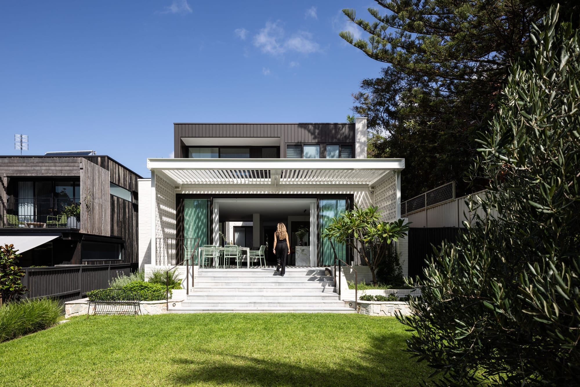 Rear view of a contemporary family home with a landscaped lawn, stepped terrace and white screened pergola opening onto bright indoor living spaces.