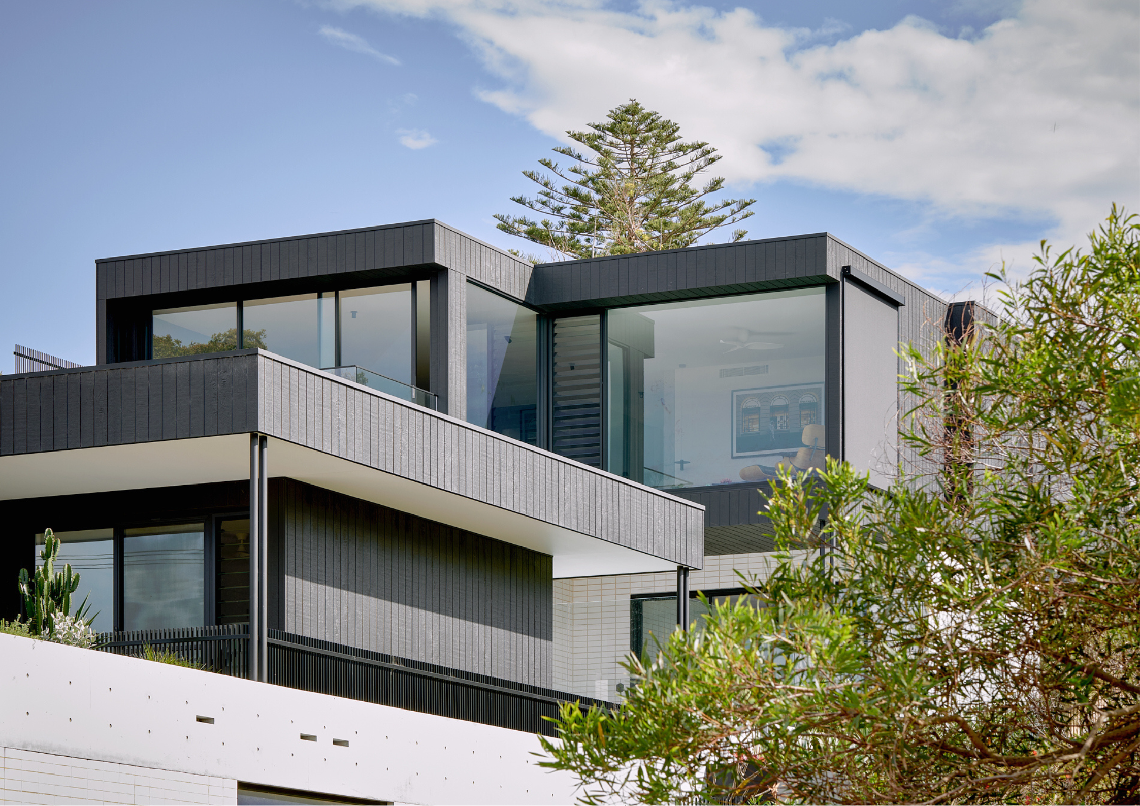 Contemporary multi-storey home with dark cladding, expansive glazing, and layered geometric forms framed by coastal vegetation.