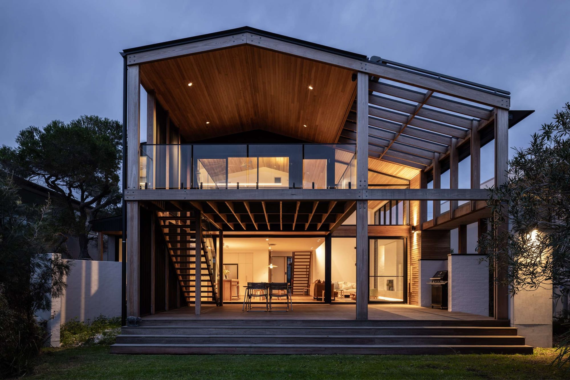 Elevated timber-framed coastal home with vaulted rooflines, open-air entertaining spaces, and warm interior lighting at dusk.