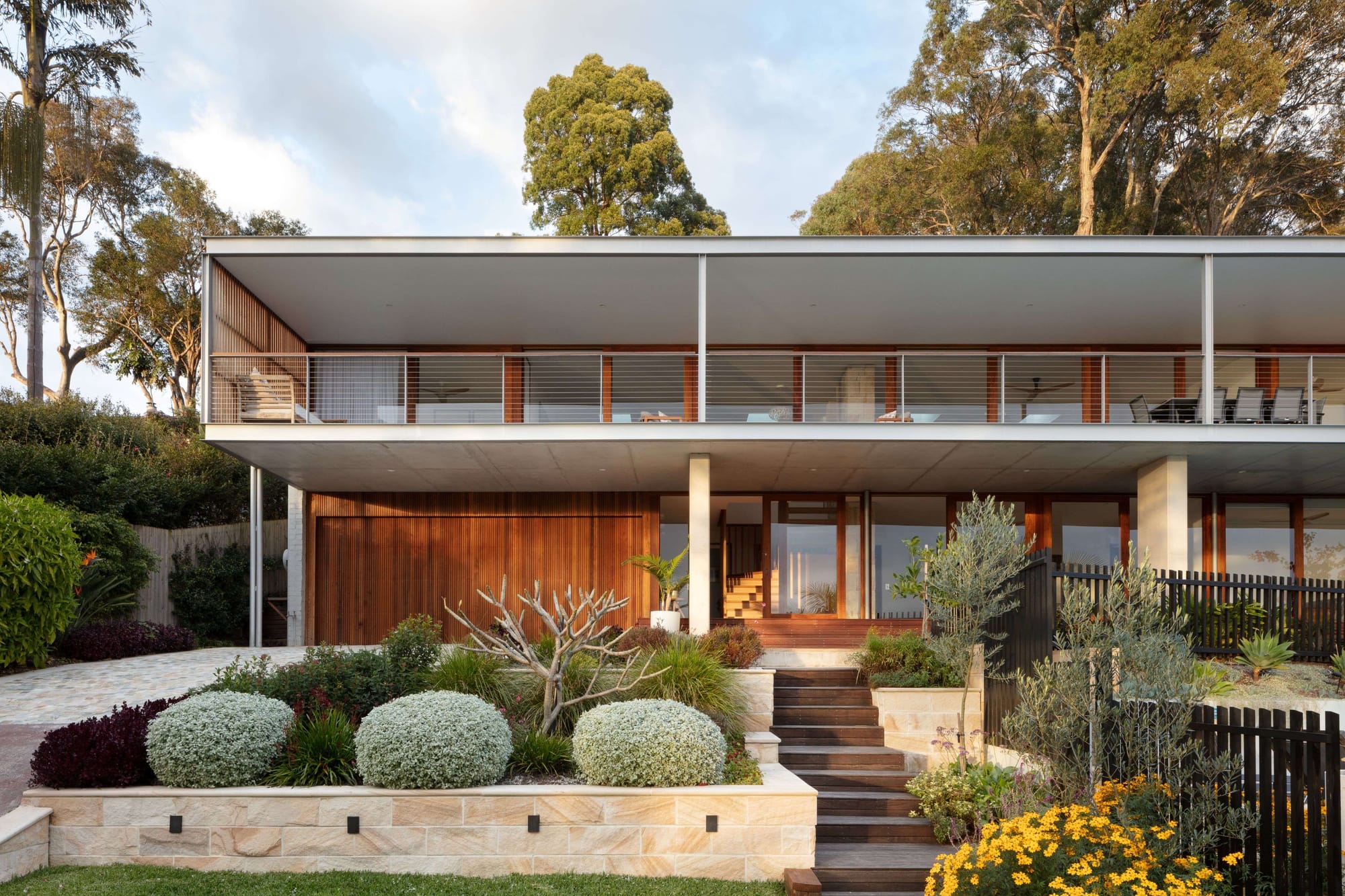 Contemporary two-storey home with expansive upper-level balcony, timber detailing, and landscaped native gardens set within a leafy bushland setting.