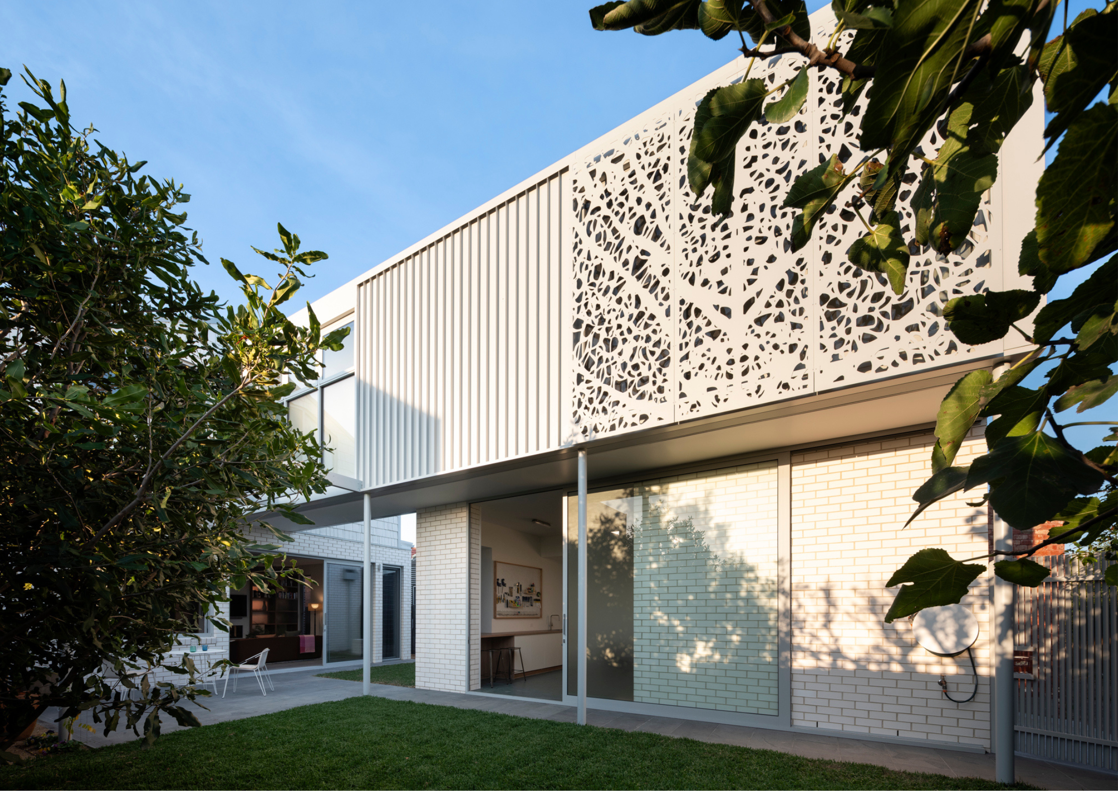 Modern two-storey home with white brick walls, large sliding glass doors, and decorative perforated metal screening on the upper level, surrounded by lush greenery and a landscaped courtyard.