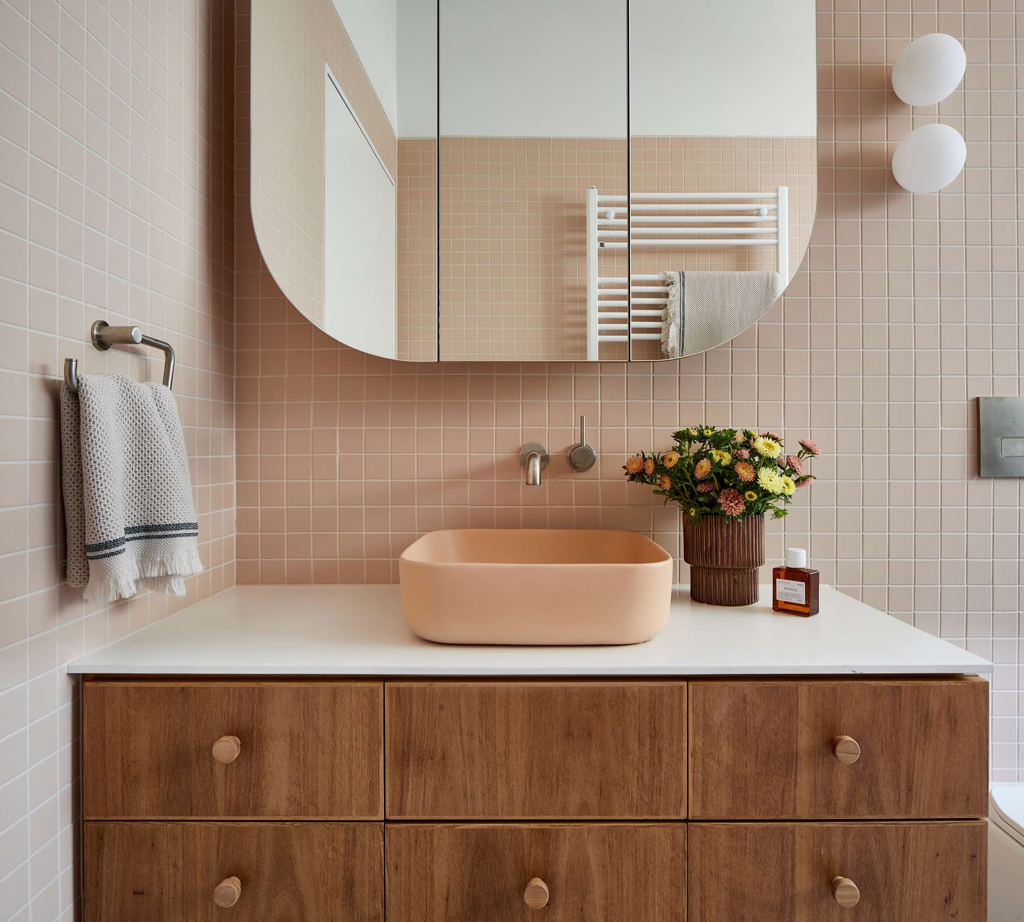 Soft pink tiled bathroom vanity with timber cabinetry, rounded mirrored cabinet and pastel basin styling.