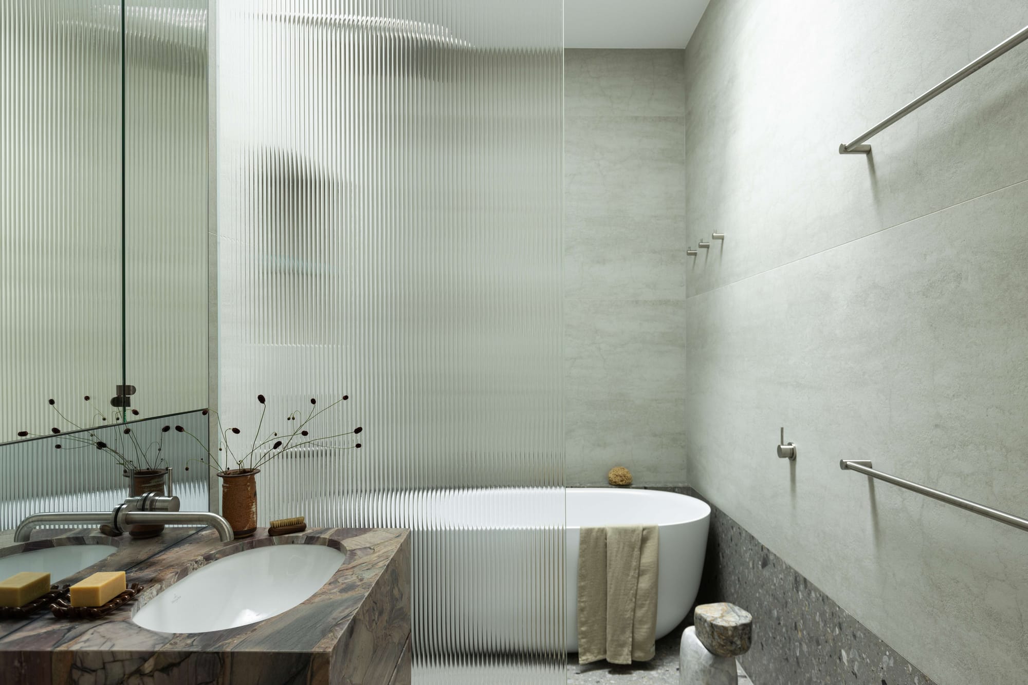 Minimal bathroom with textured stone surfaces, ribbed glass partition and sculptural basin with muted neutral tones.
