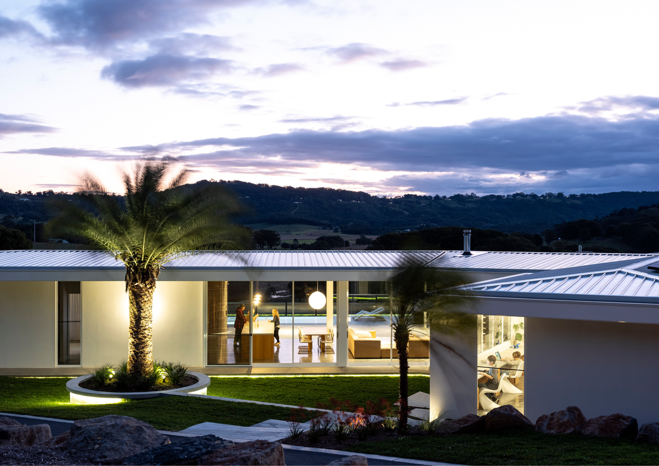 Minimal contemporary pavilion home with expansive glazing and illuminated interiors overlooking rolling rural hills at dusk.