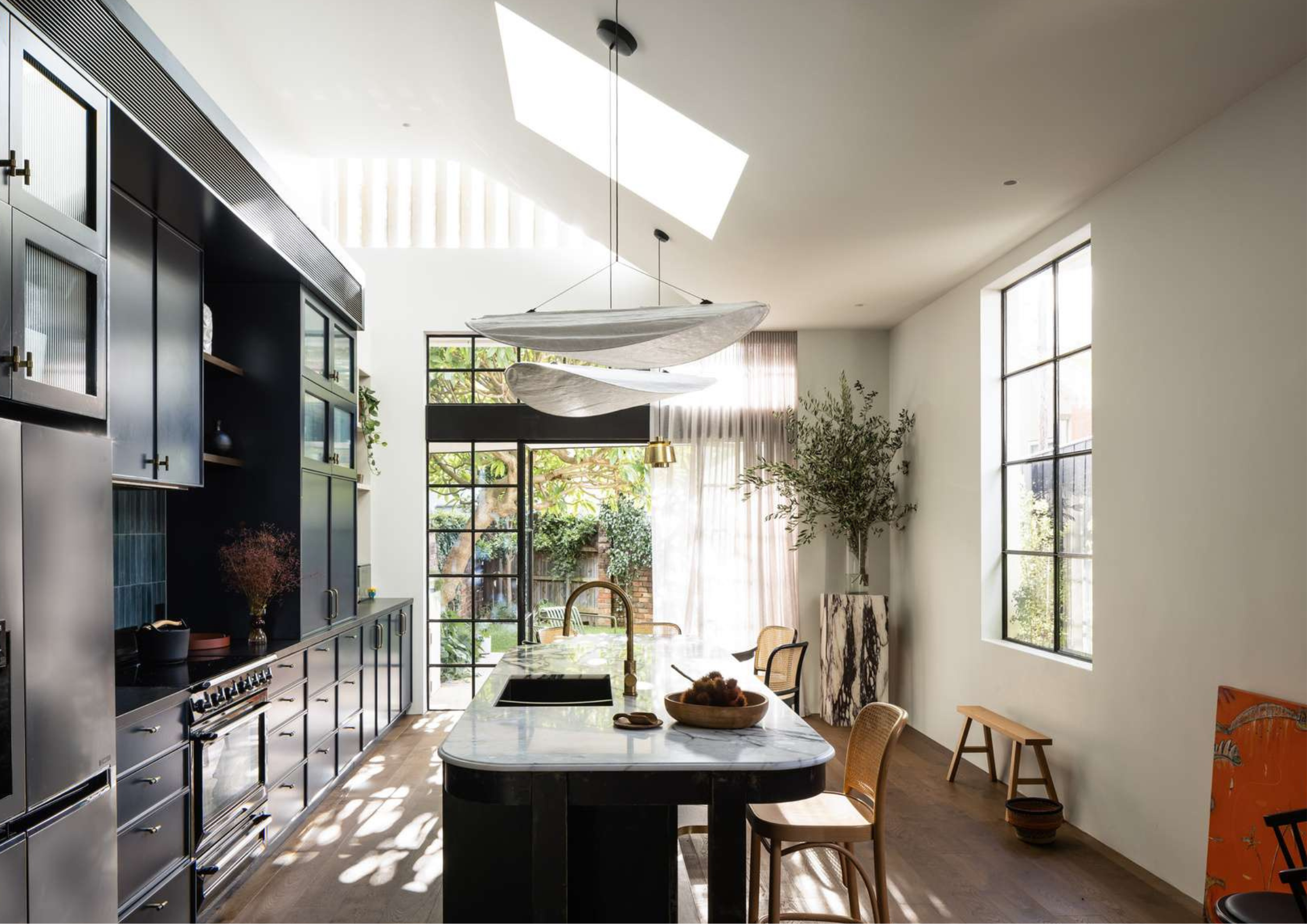 Light-filled galley kitchen with dark joinery, marble island bench, skylights, and sculptural pendant lighting.