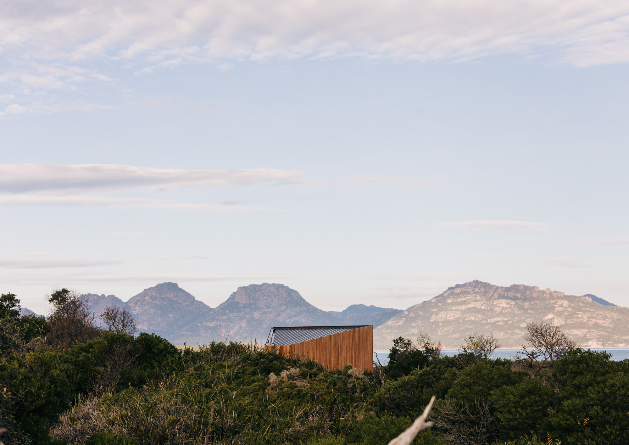 Small timber-clad studio nestled within coastal bushland, framed by distant mountain ranges beneath a soft pastel sky.