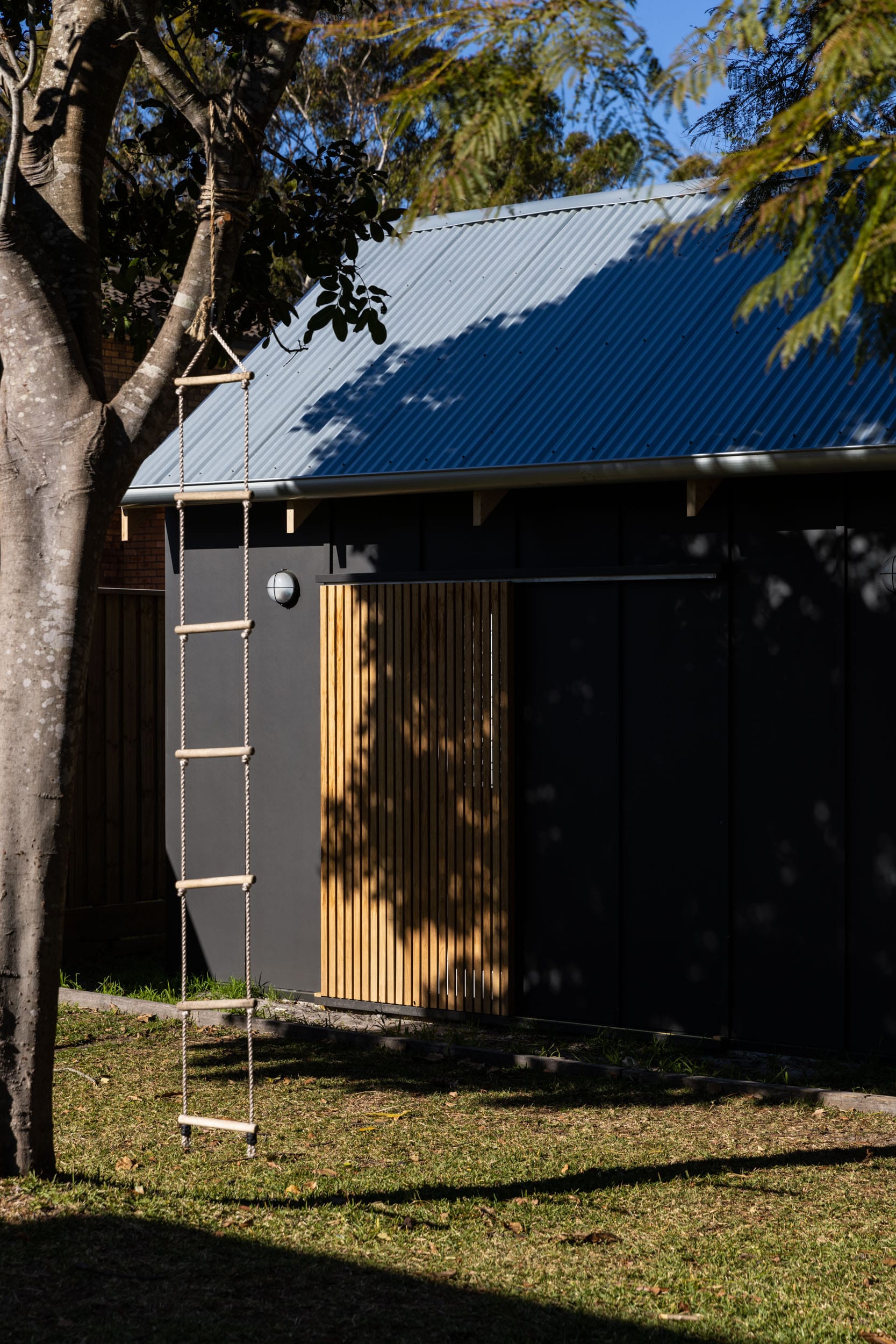 Black-clad outdoor structure with corrugated roofing and timber detailing set within a backyard garden.
