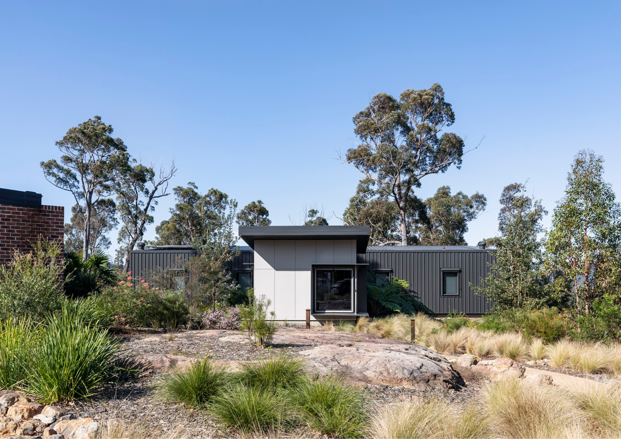 Single-storey contemporary home with dark metal cladding surrounded by native planting and rocky natural terrain.