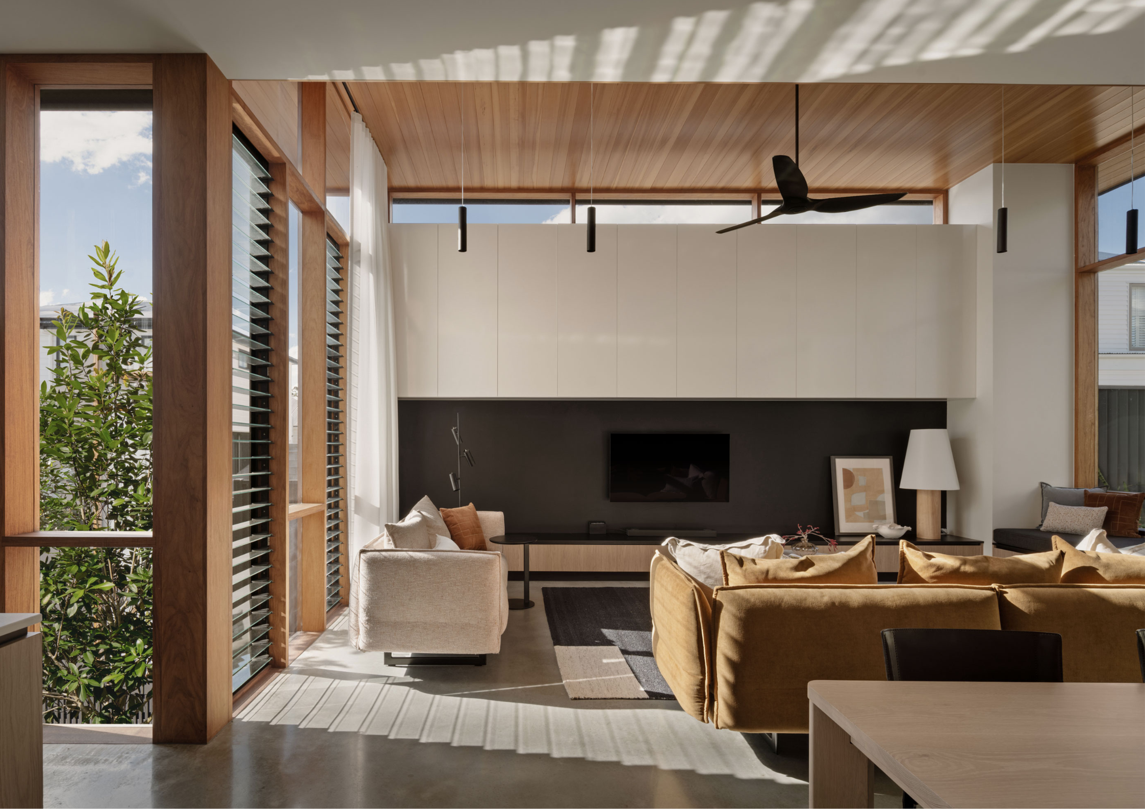 Sunlit contemporary living room with timber-lined ceilings, expansive glazing, and warm neutral furnishings opening to a subtropical garden outlook.
