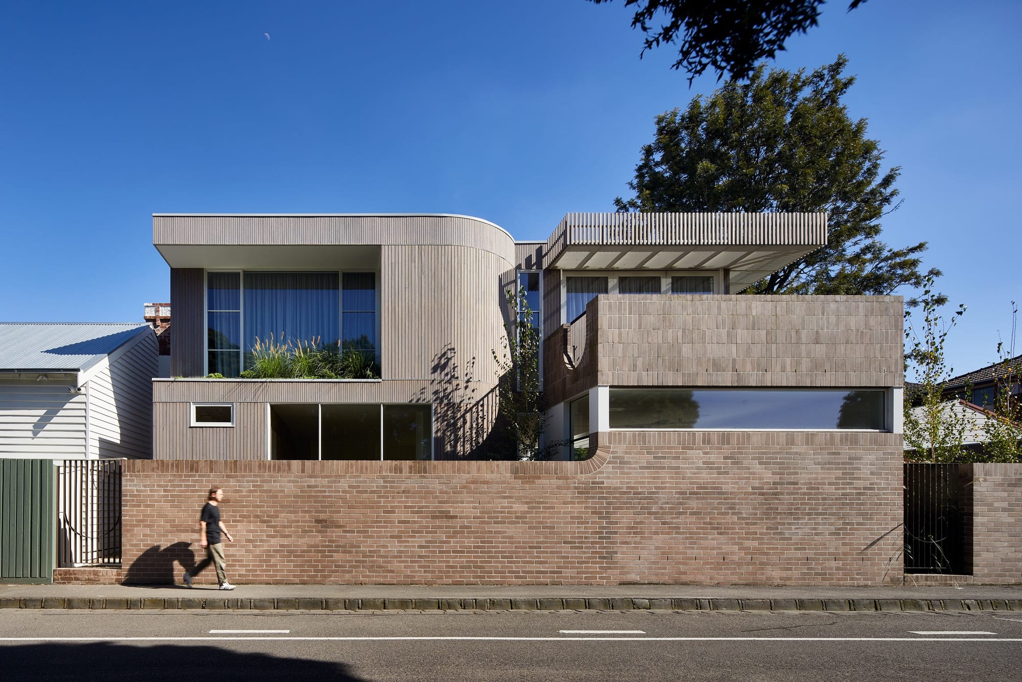 Modern residence featuring curved timber cladding, textured brickwork, and screened upper terraces behind a long masonry boundary wall.