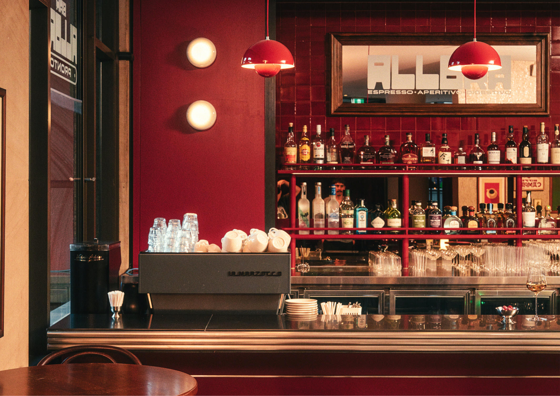 Deep red bar interior with glossy tiled walls, red pendant lights and mirrored shelving stocked with bottles and glassware behind a stainless steel counter.