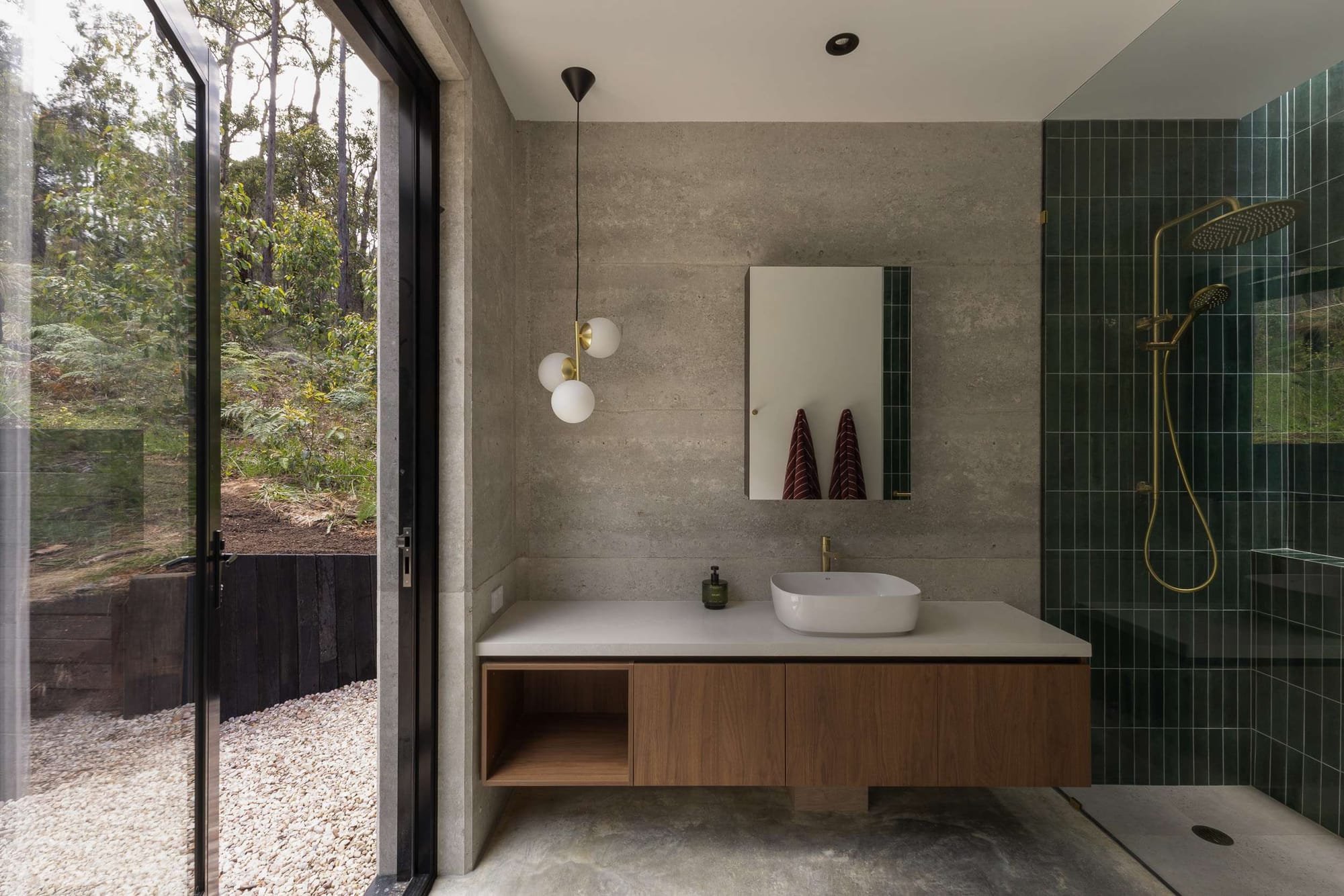 Contemporary bathroom with concrete walls, timber vanity and dark green tiled shower overlooking a natural bushland setting.