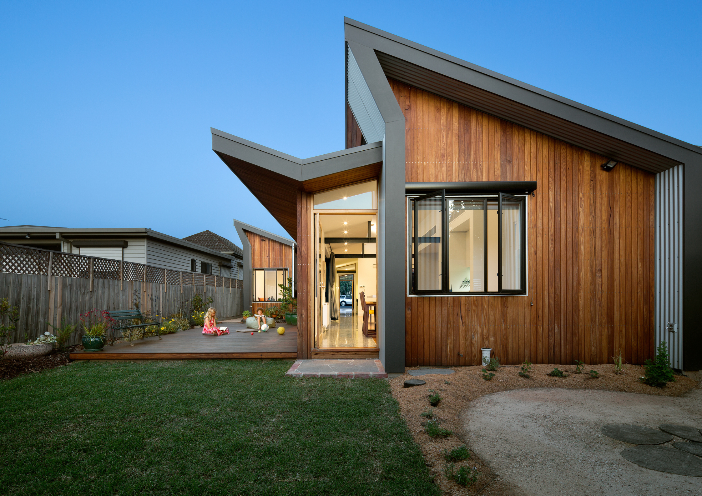 Compact timber-clad house with angular roof opening to a backyard deck and garden.
