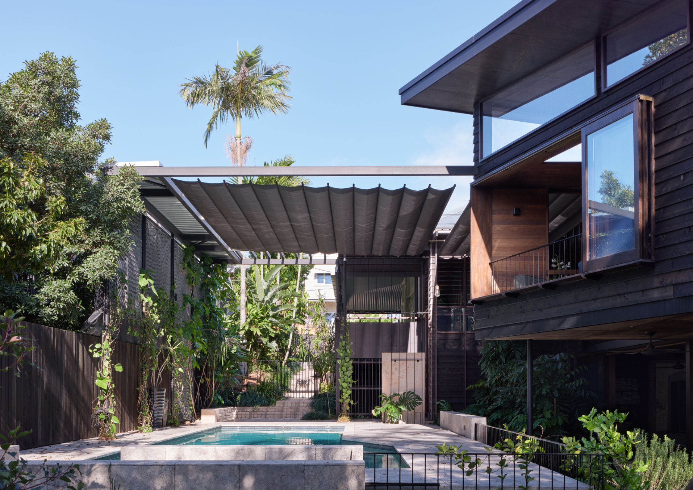 Tropical-inspired courtyard with swimming pool, retractable shade canopy, and dark timber-clad architecture surrounded by dense planting.