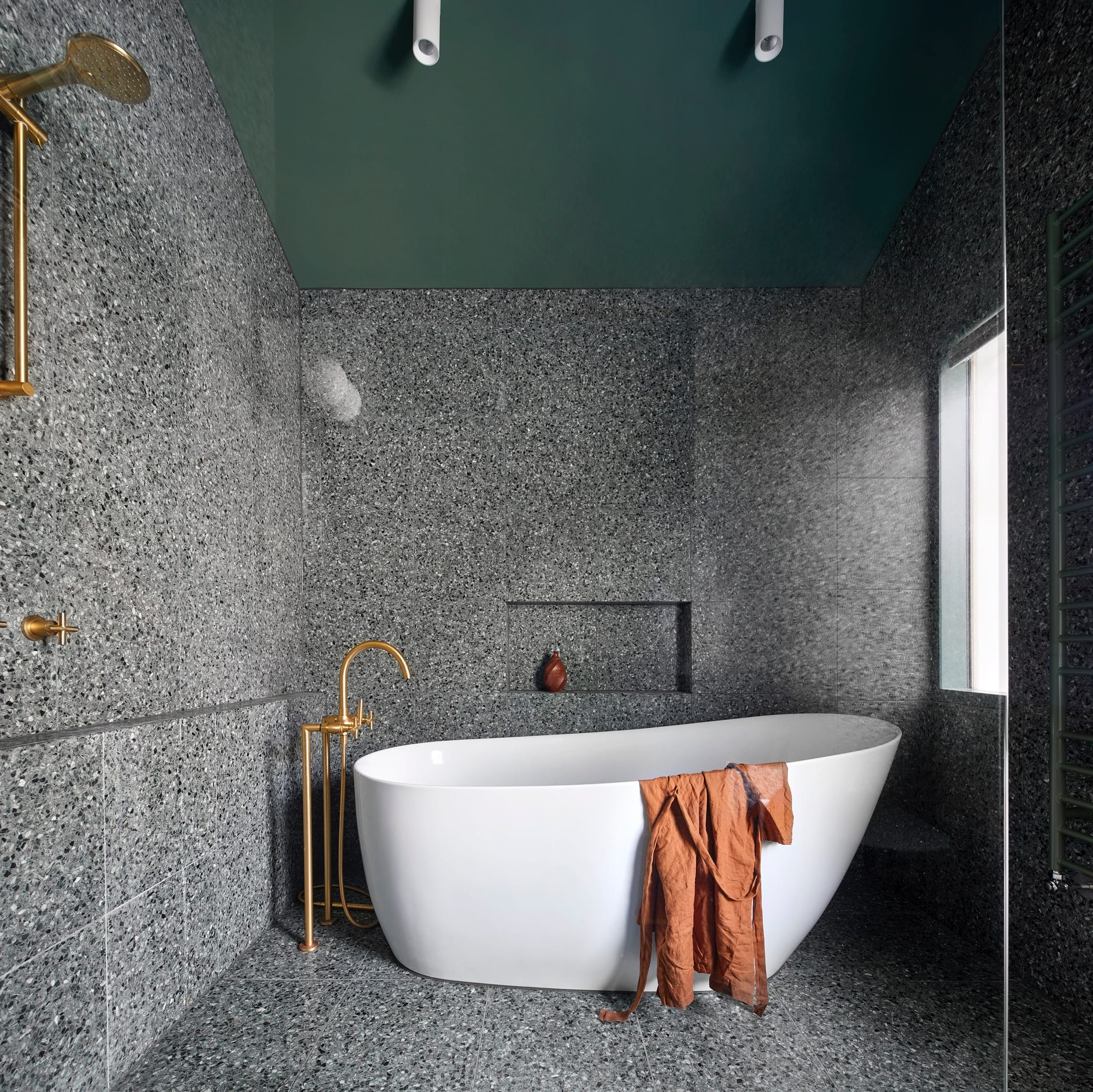 Speckled stone bathroom with freestanding bath, brass fixtures and deep green ceiling creating a dramatic mood.