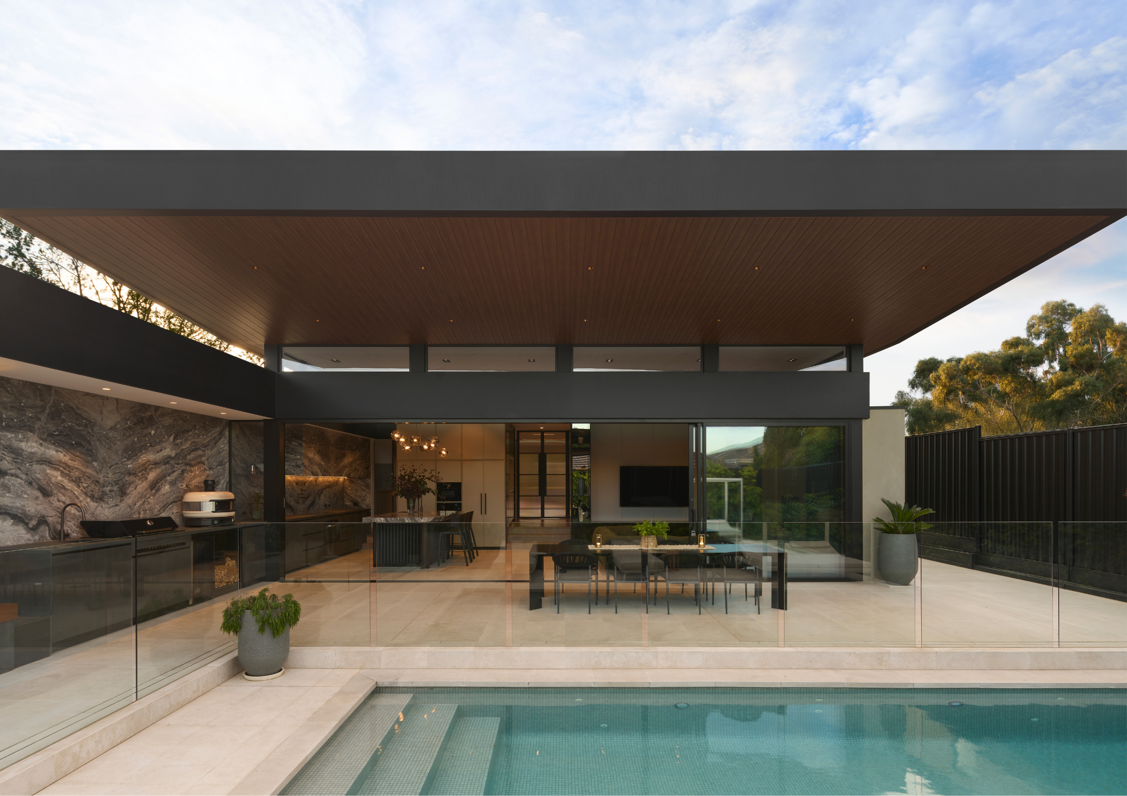Contemporary outdoor kitchen and dining area beneath a wide timber-lined roof beside a pool.