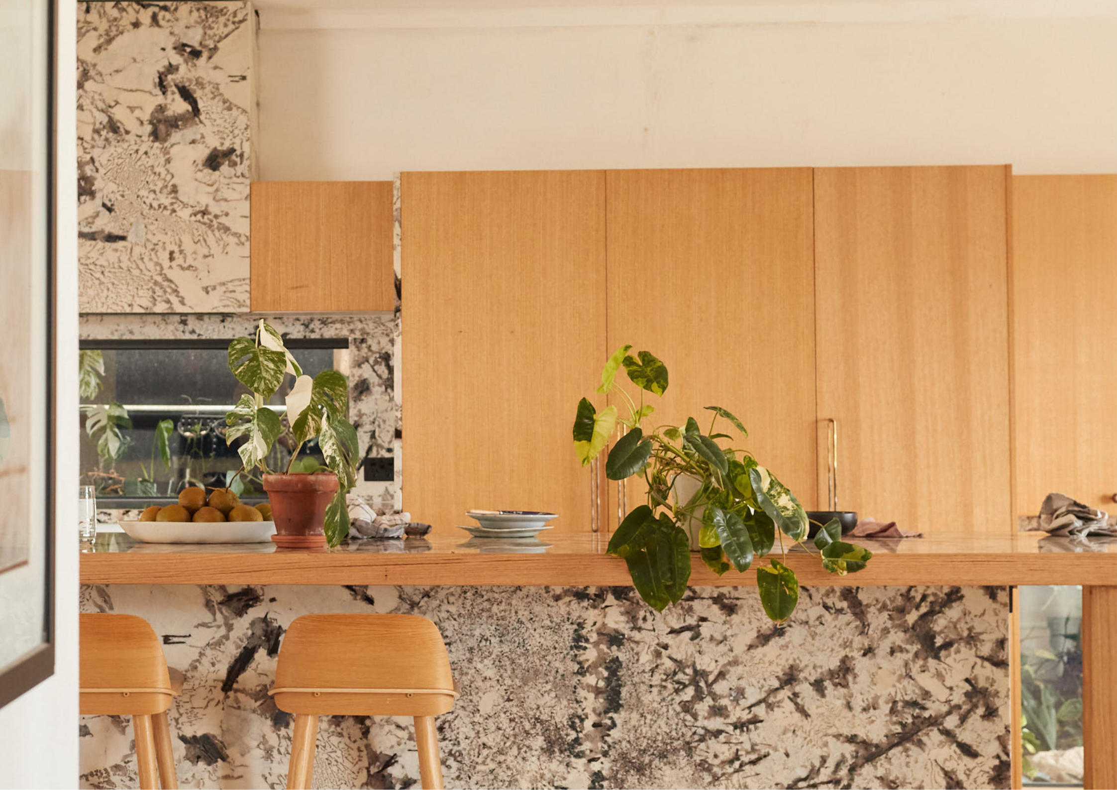 Warm timber kitchen with light wood cabinetry, a marble island with expressive veining, indoor plants and simple timber stools in soft natural light.