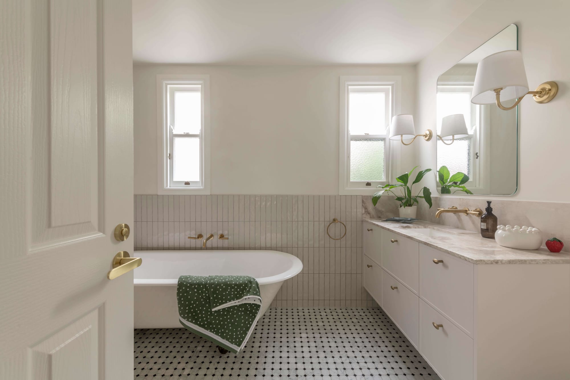 Light-filled bathroom with marble vanity, brass fixtures and freestanding bathtub against vertically stacked white tiles.