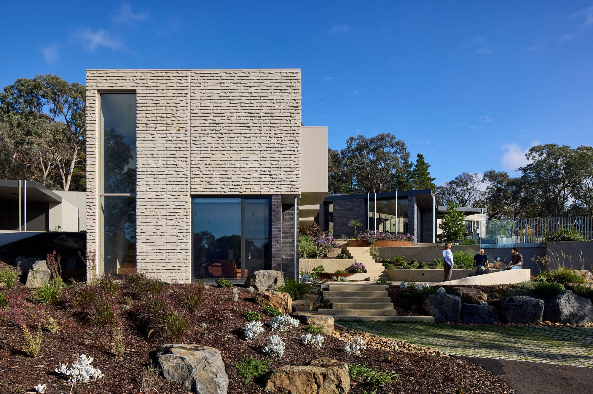 Modern courtyard framed by angled rooflines, full-height glazing, and natural stone paving surrounded by native planting.