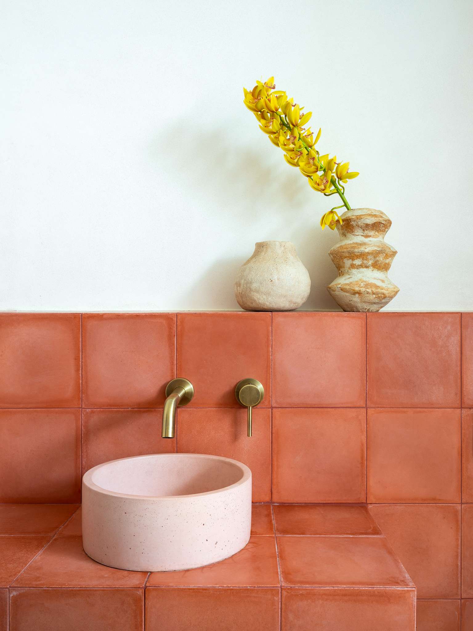 Terracotta tiled vanity with round stone basin, brass tapware and ceramic vessels with yellow orchid arrangement.