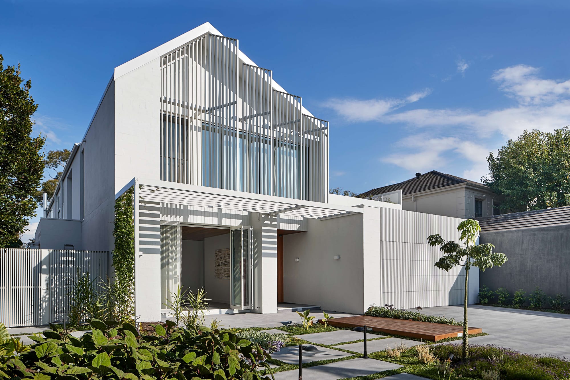 Minimalist white residence with sculptural vertical screening, clean geometric lines, and landscaped front garden under a bright blue sky.