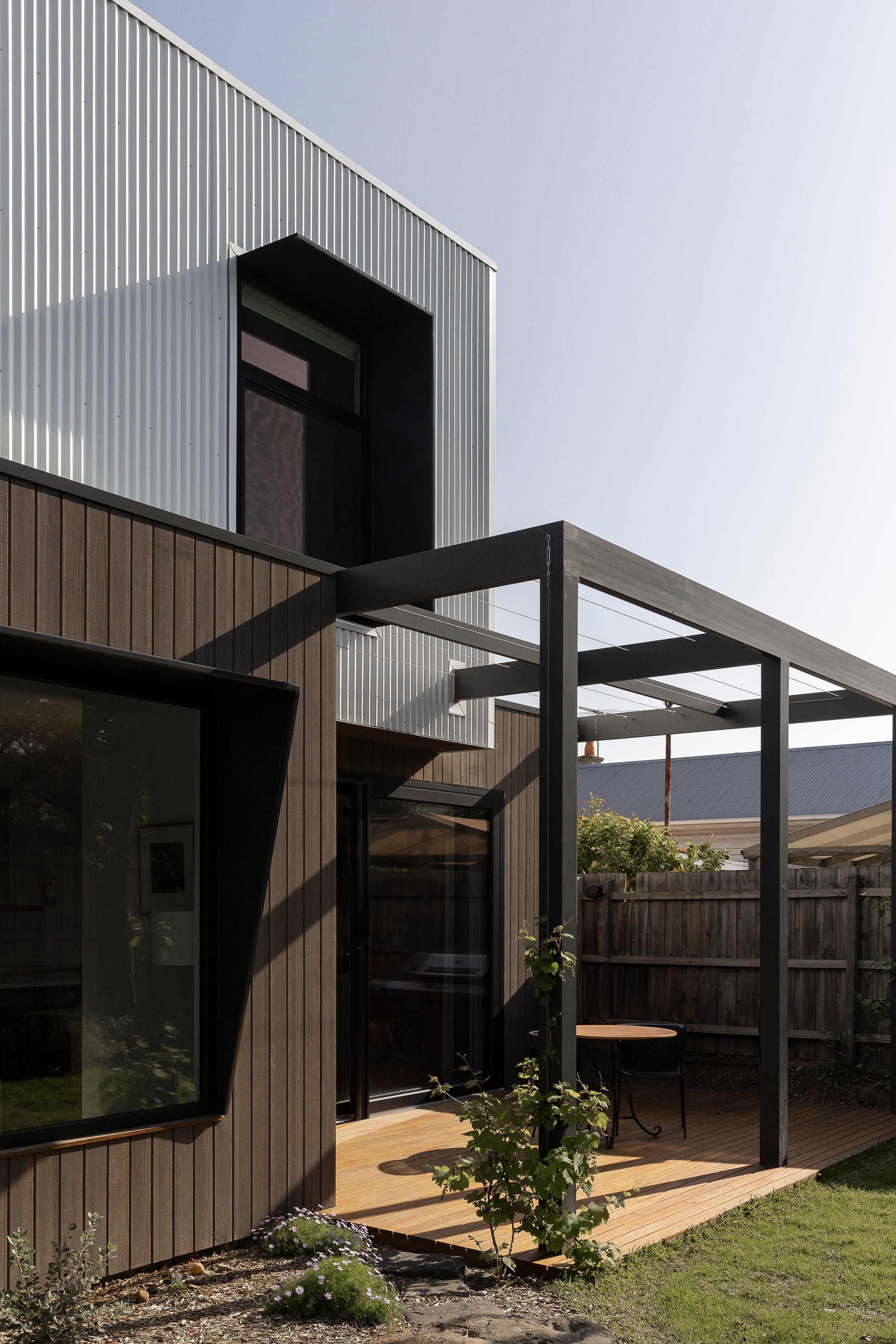 Detail view of a contemporary home featuring dark cladding, steel pergola structure, and corrugated metal upper volume.