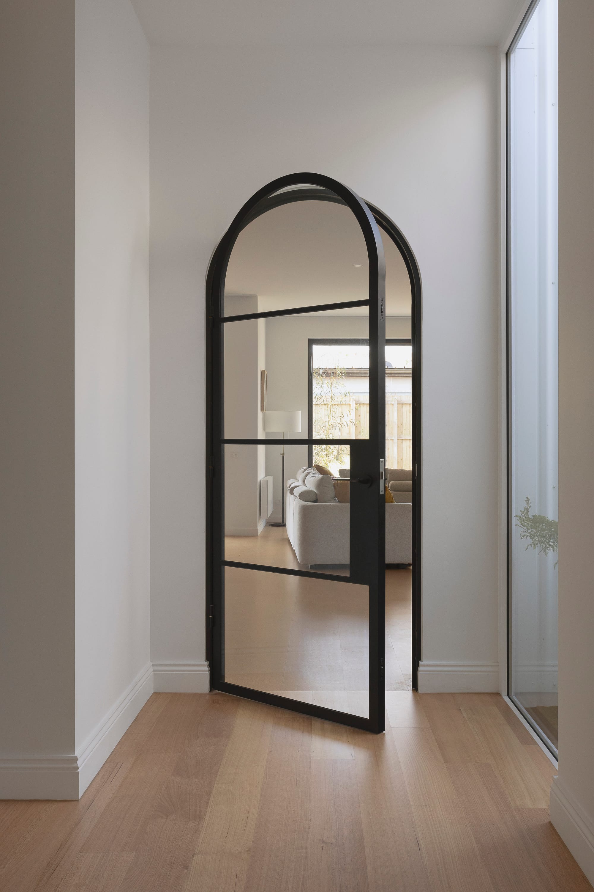 Minimal hallway featuring an arched black steel-framed glass door, light timber flooring, and soft natural light leading into a contemporary living space.