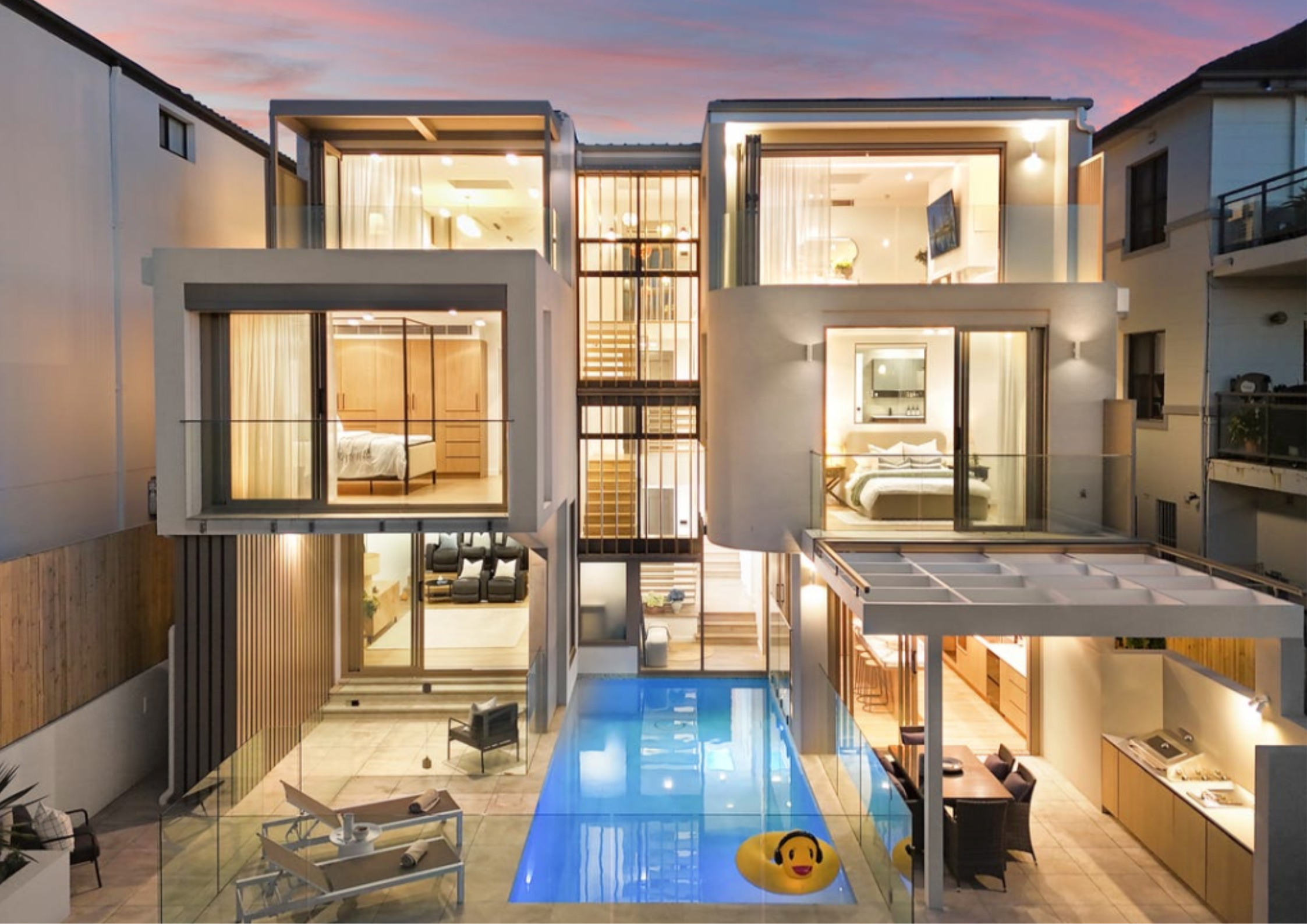 Modern multi-level home with projecting box volumes, expansive glazing, central stair void, and swimming pool courtyard illuminated at dusk.