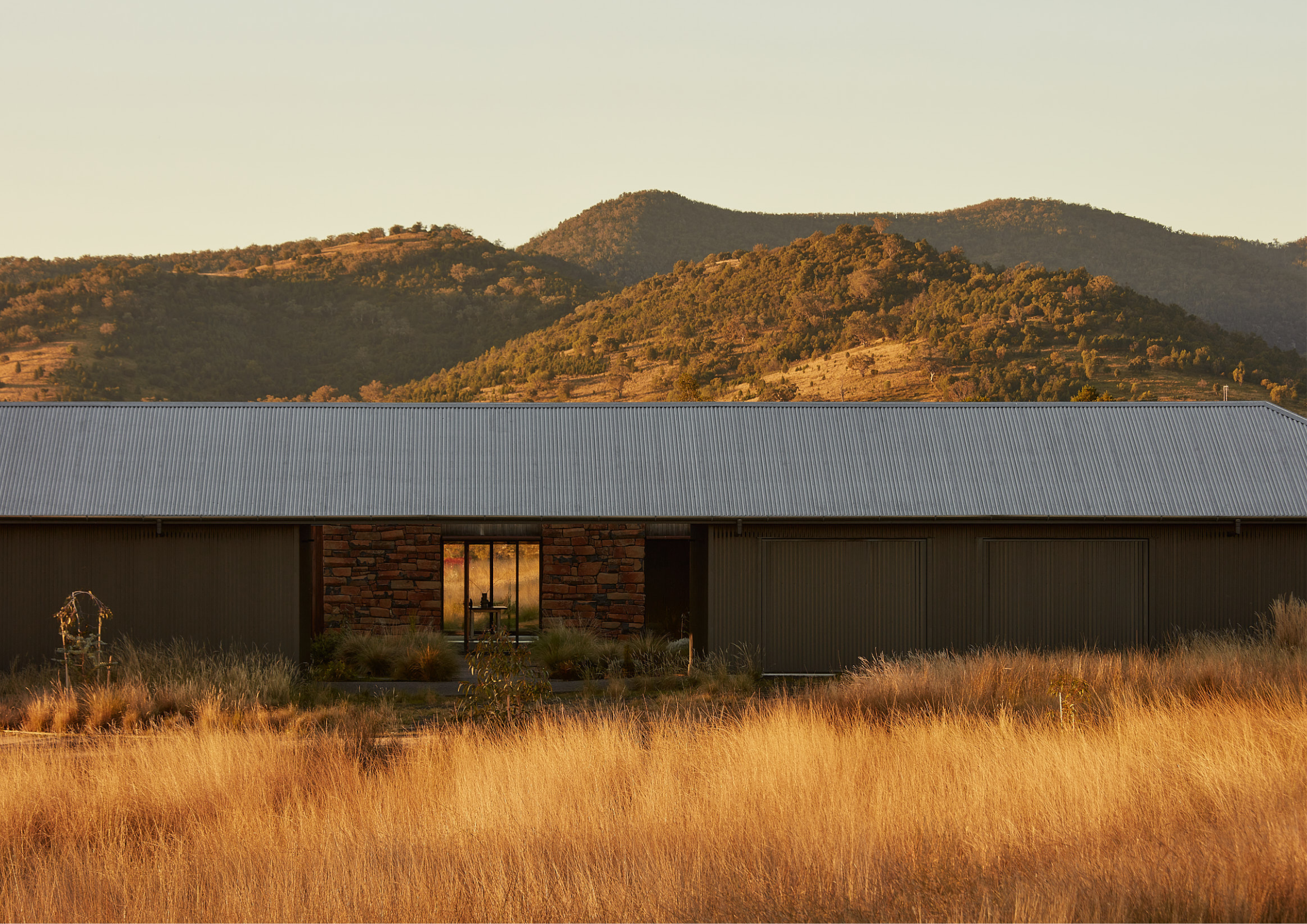 Rural house with corrugated roof and stone walls set against dry grassy landscape and hills.