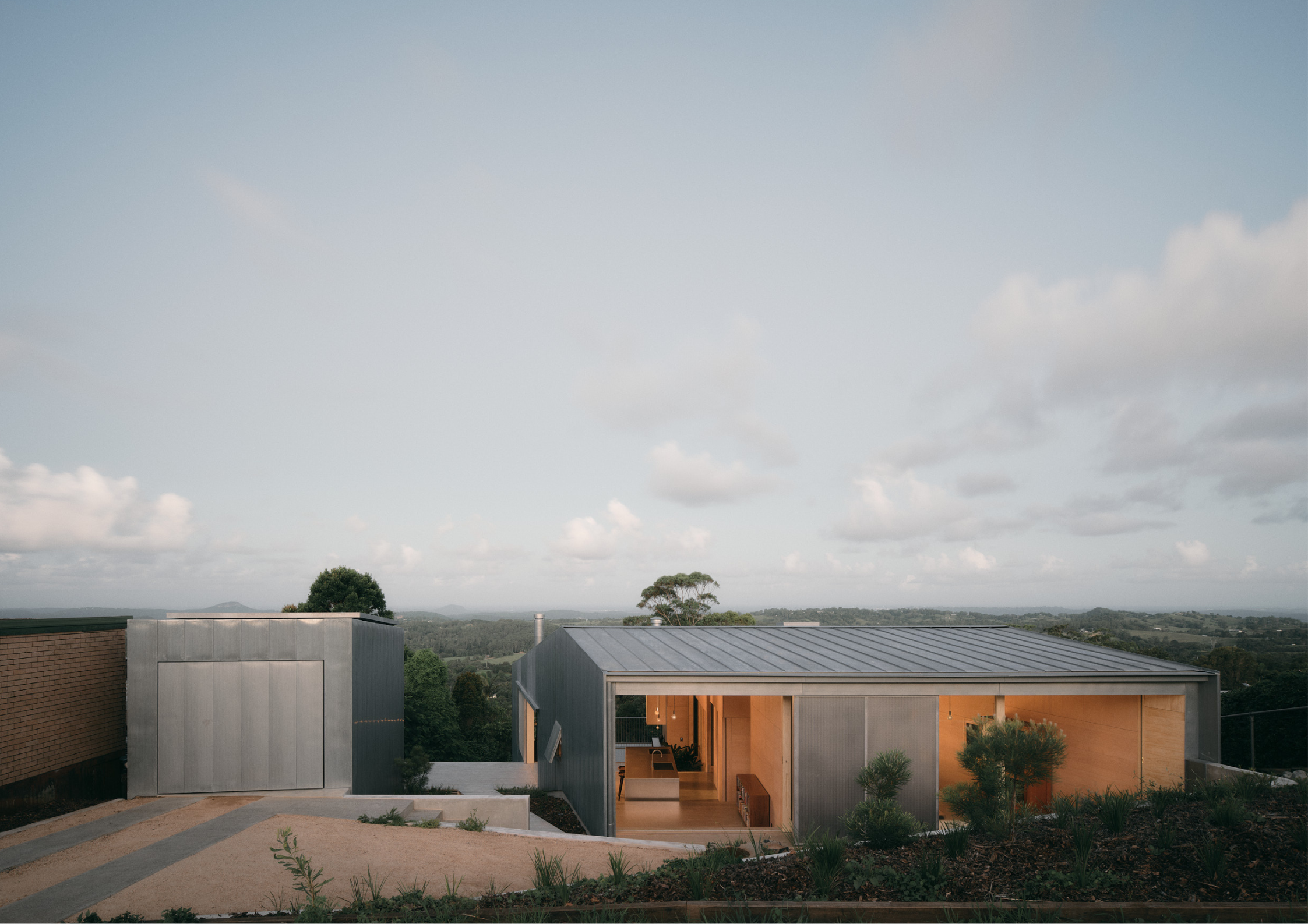 Minimal hillside home with corrugated metal cladding and gabled forms overlooking a lush rural landscape at dusk.