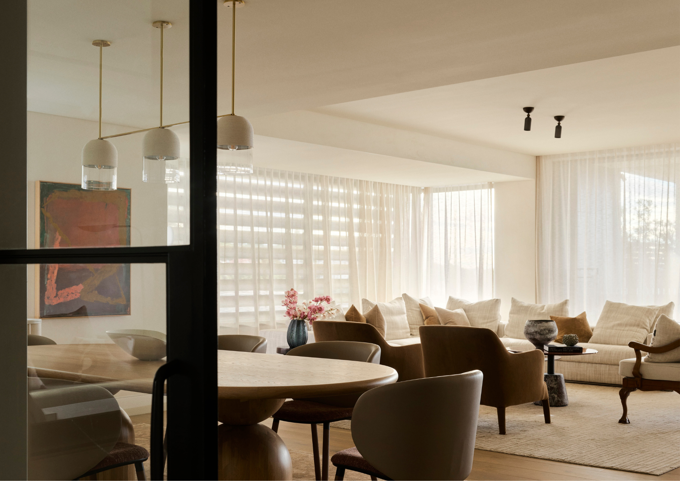 Softly lit open-plan living and dining space with sheer curtains, curved upholstered seating, warm timber furniture and layered neutral textures.