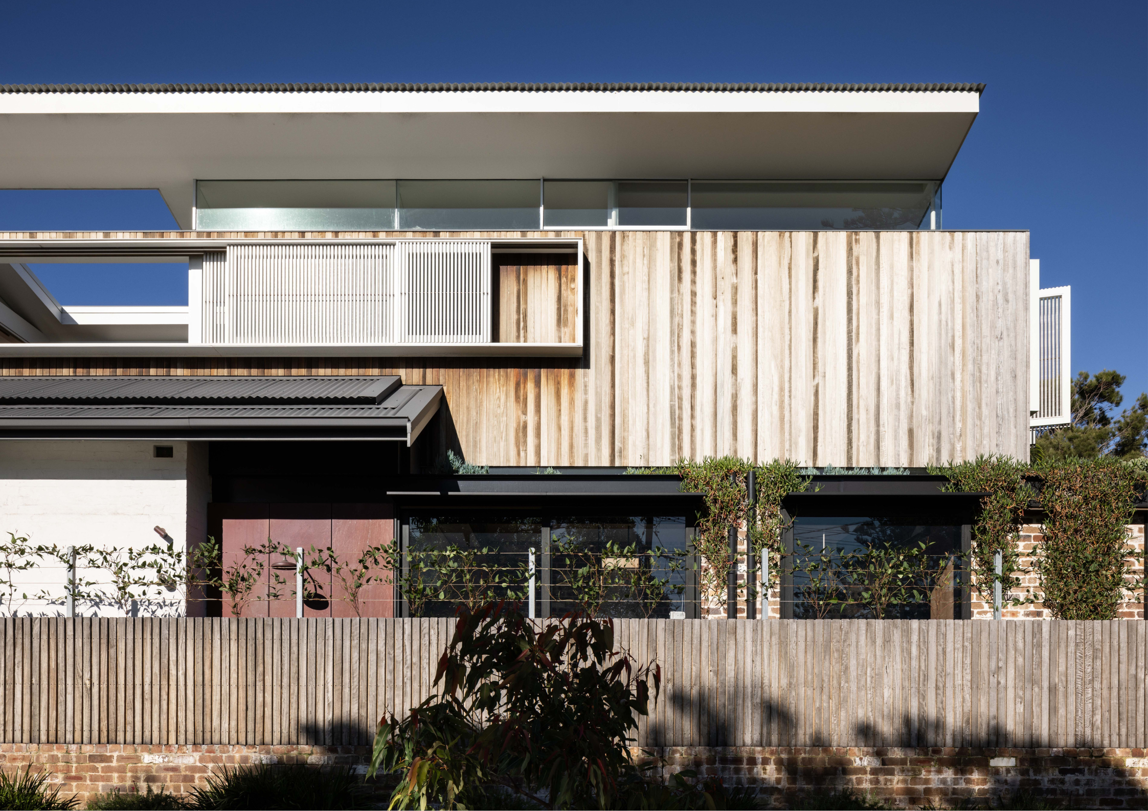 Contemporary coastal-style residence with timber battens, white-framed balcony screens, and layered horizontal rooflines above a landscaped frontage.