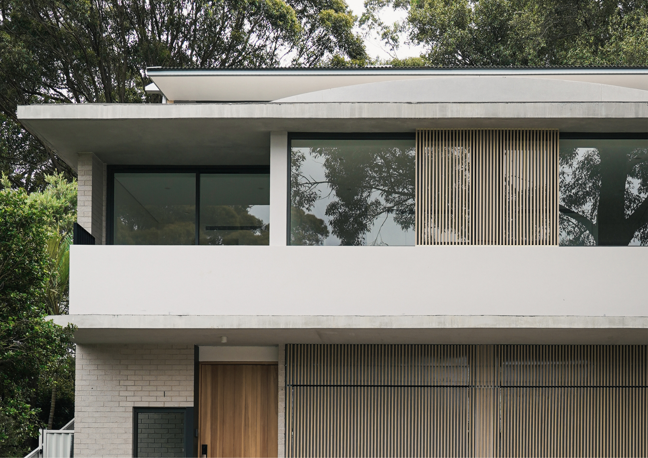 Minimalist two-storey house with white concrete slabs, vertical timber screening, and large recessed windows framed by surrounding trees.
