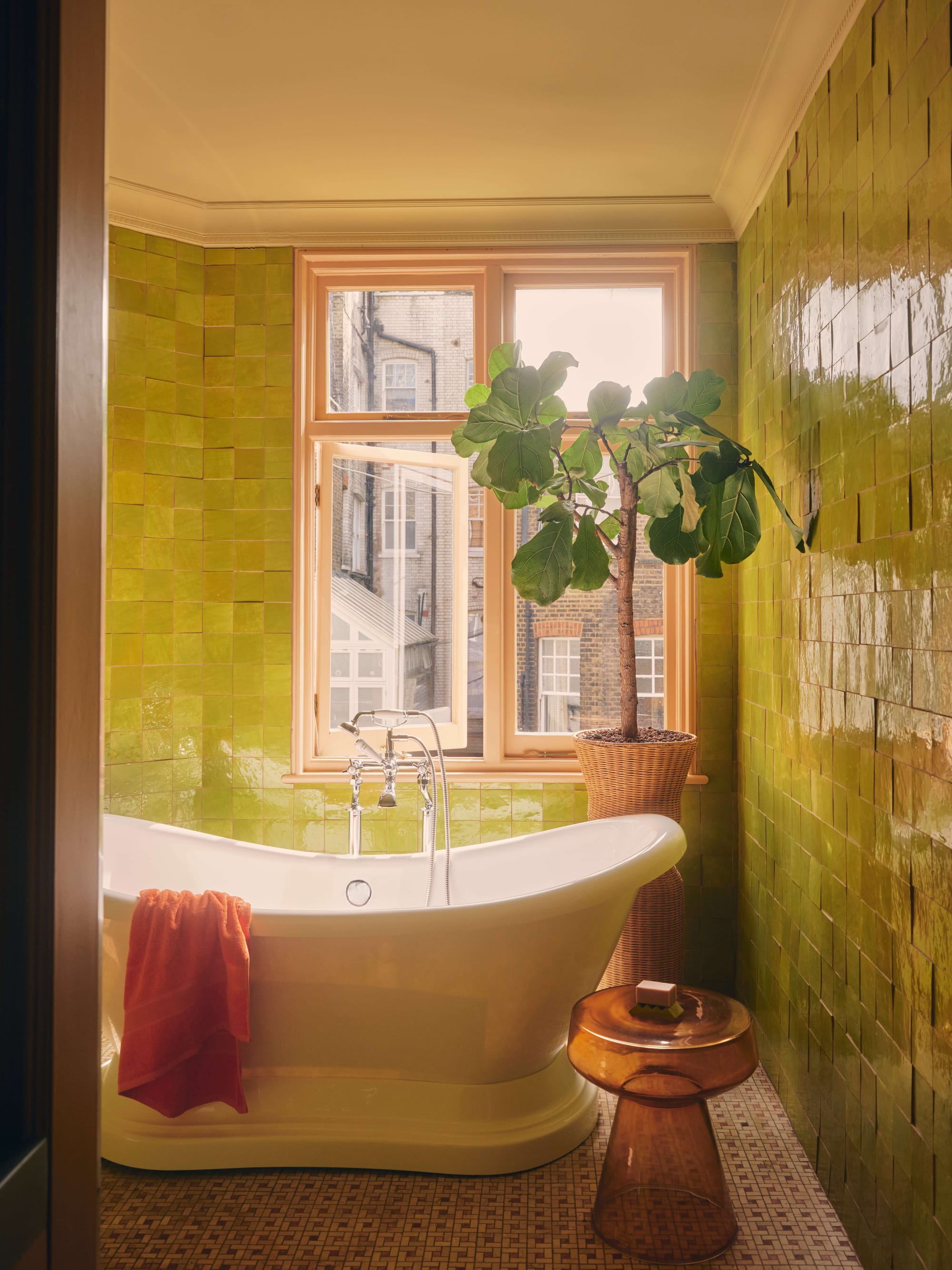 Warm bathroom interior with glossy olive-green wall tiles, freestanding bathtub and large window with indoor plant.