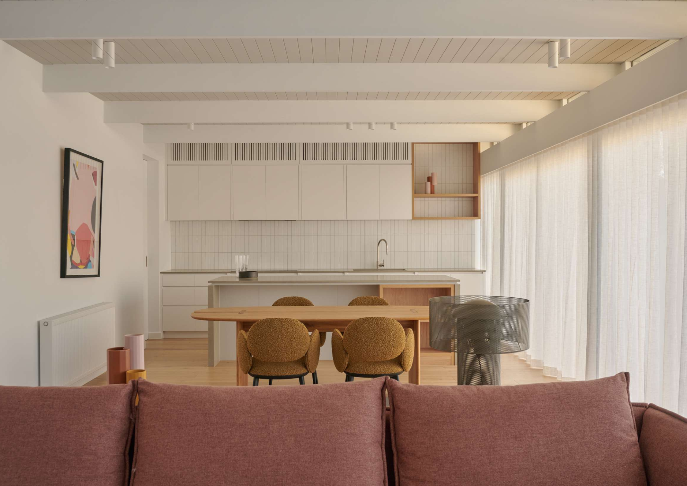 Minimal white kitchen with timber accents, integrated dining area, and sheer-curtained natural light.