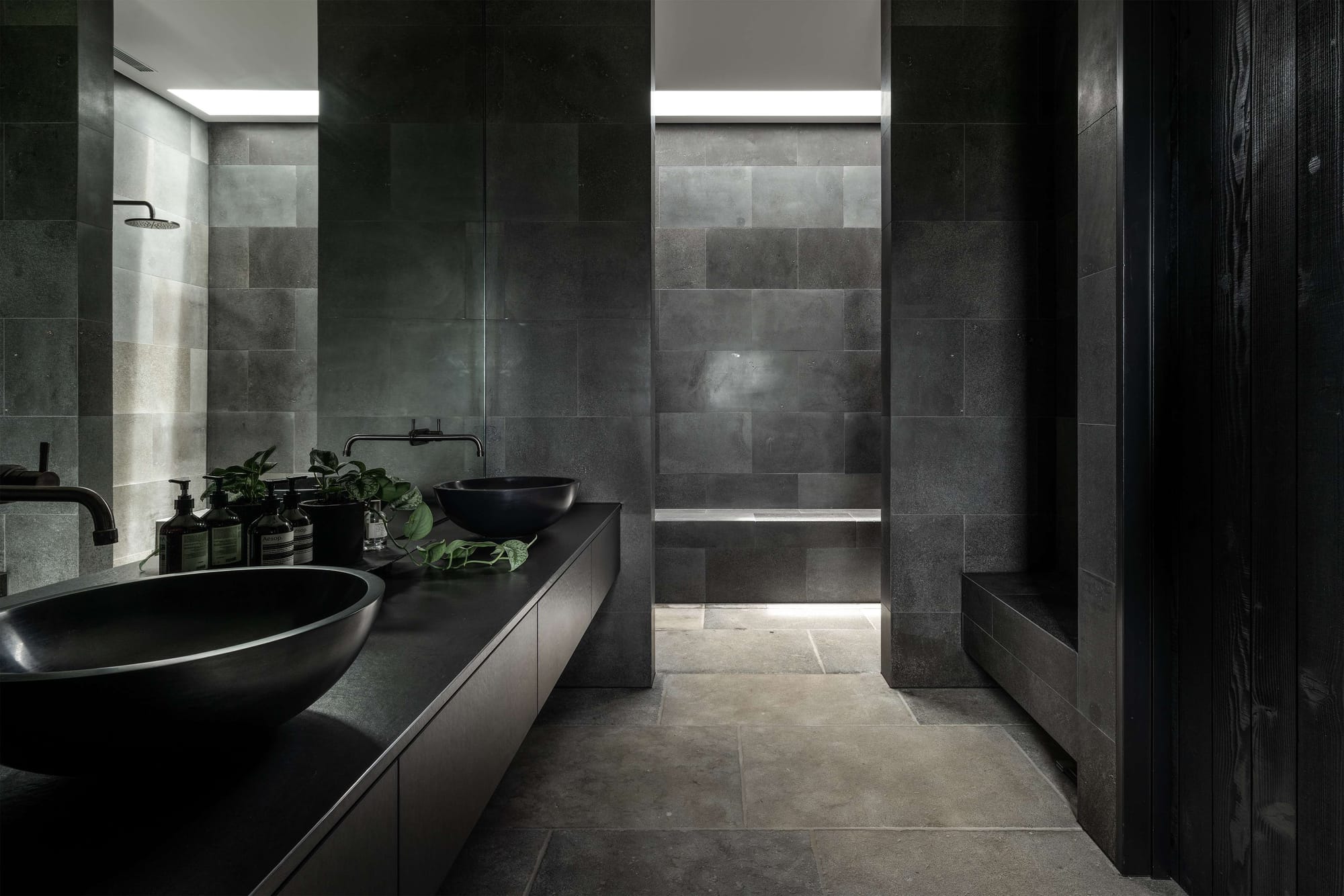 Dark stone bathroom with black basins, dramatic lighting and monolithic tiled walls creating a spa-like atmosphere.