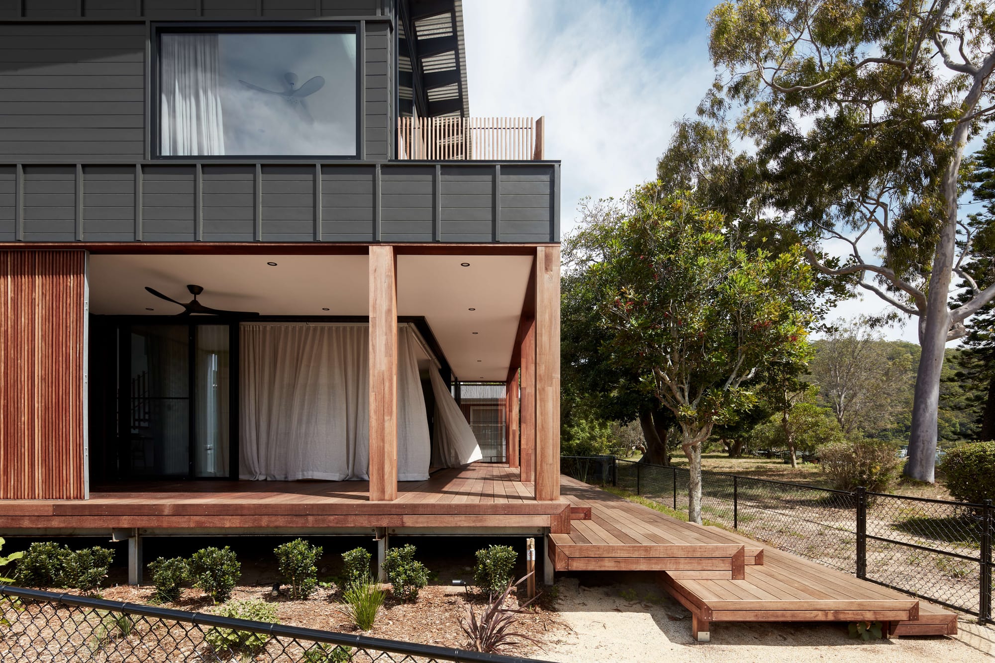 Dark-clad coastal home with timber decking, open verandah, and surrounding native landscape beside a waterfront reserve.