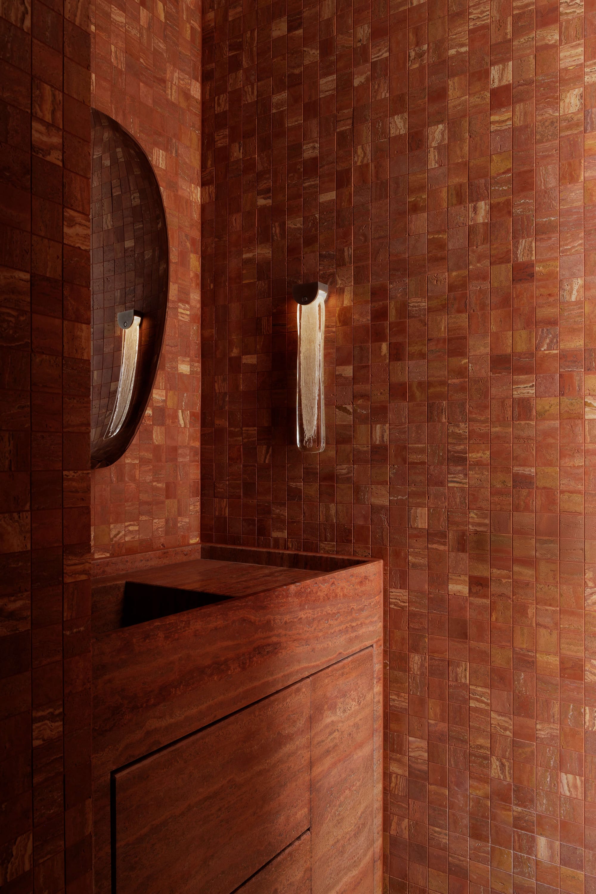 Warm, moody bathroom clad entirely in rich terracotta stone tiles, featuring a sculptural stone vanity, an oval wall mirror, and a textured glass wall light that casts a soft ambient glow.