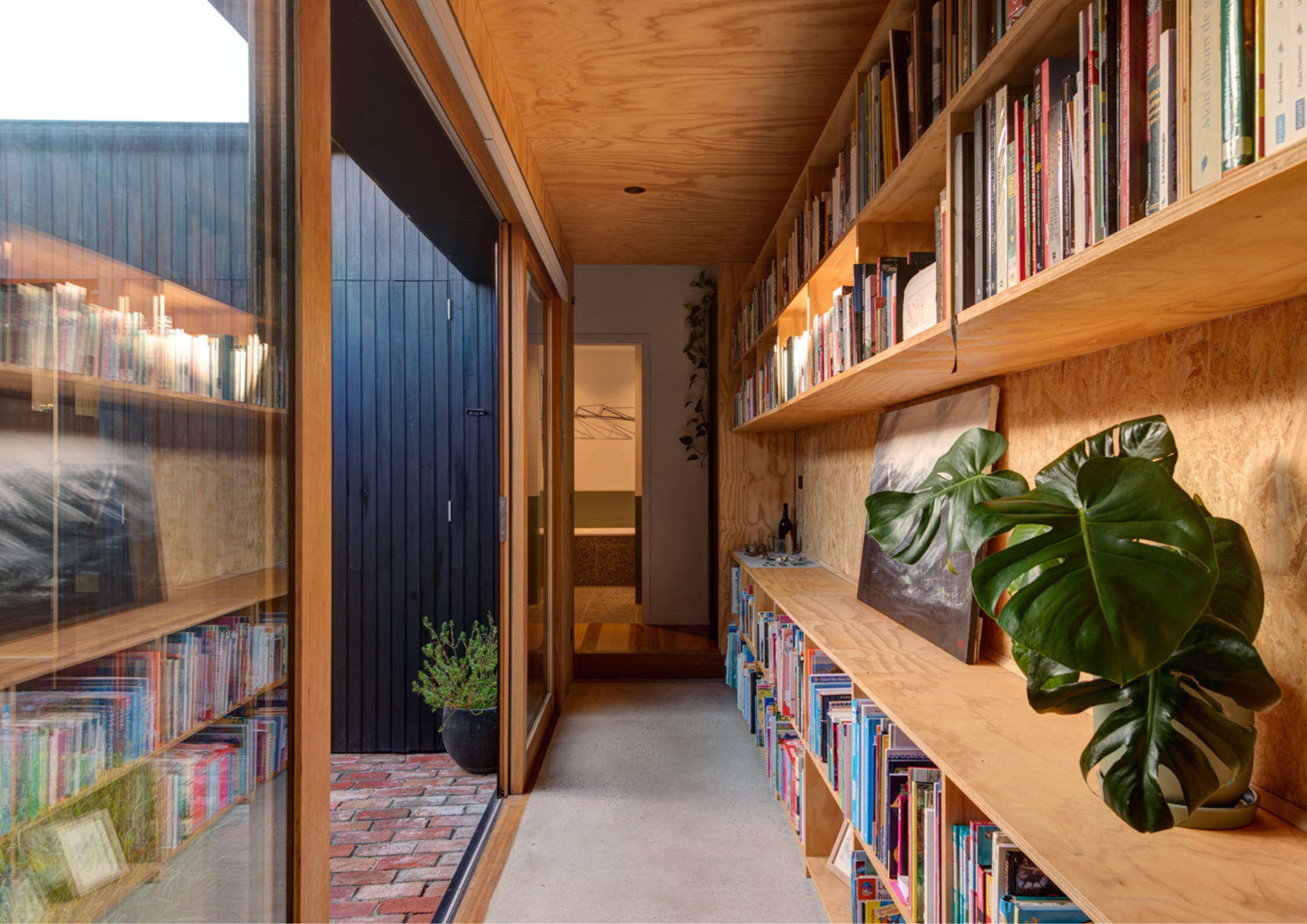 Narrow hallway lined with timber shelving filled with books and greenery, leading to a bathroom.
