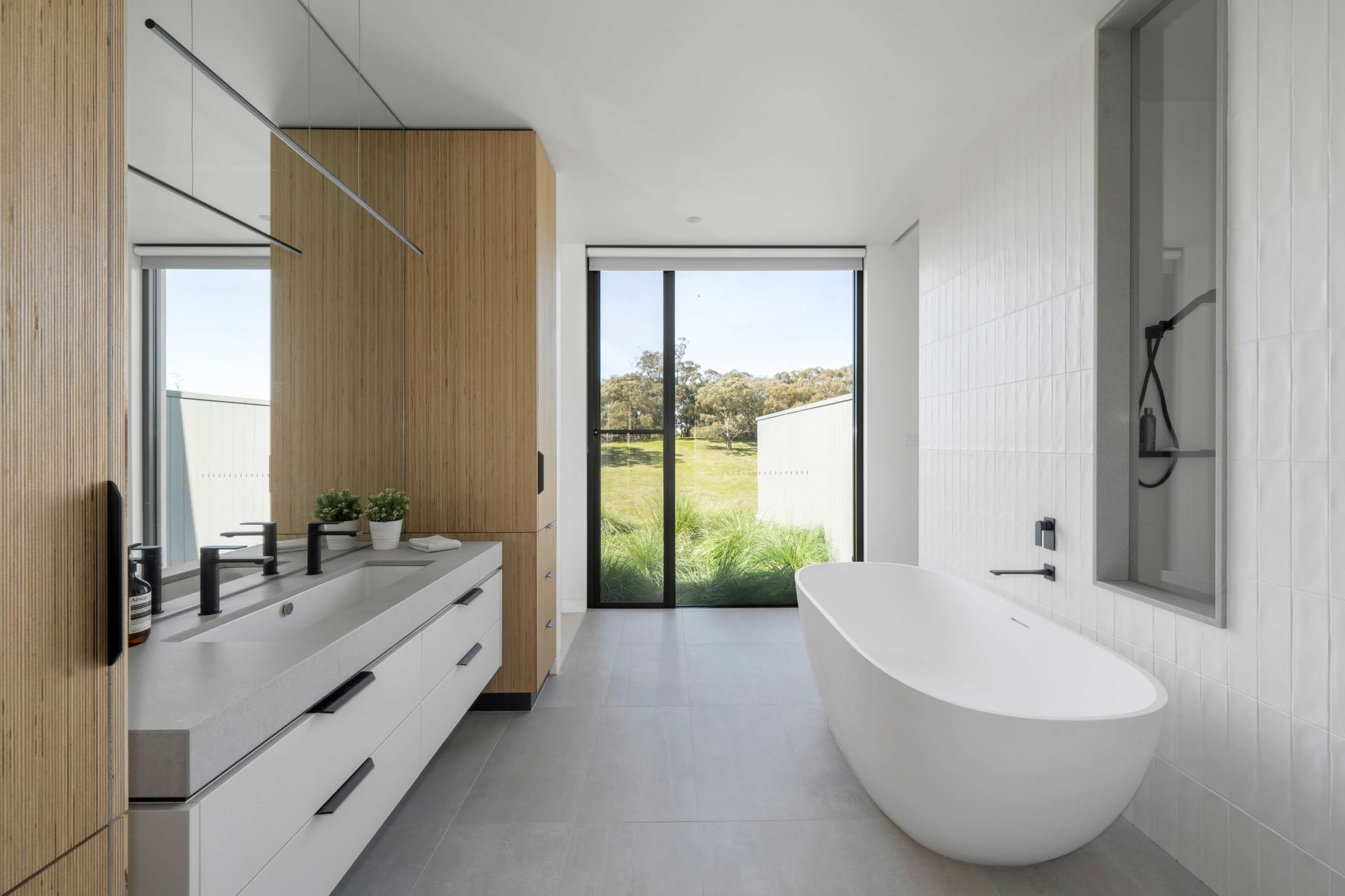 Minimal white bathroom with timber detailing, freestanding bath and floor-to-ceiling glazing overlooking a grassy landscape.