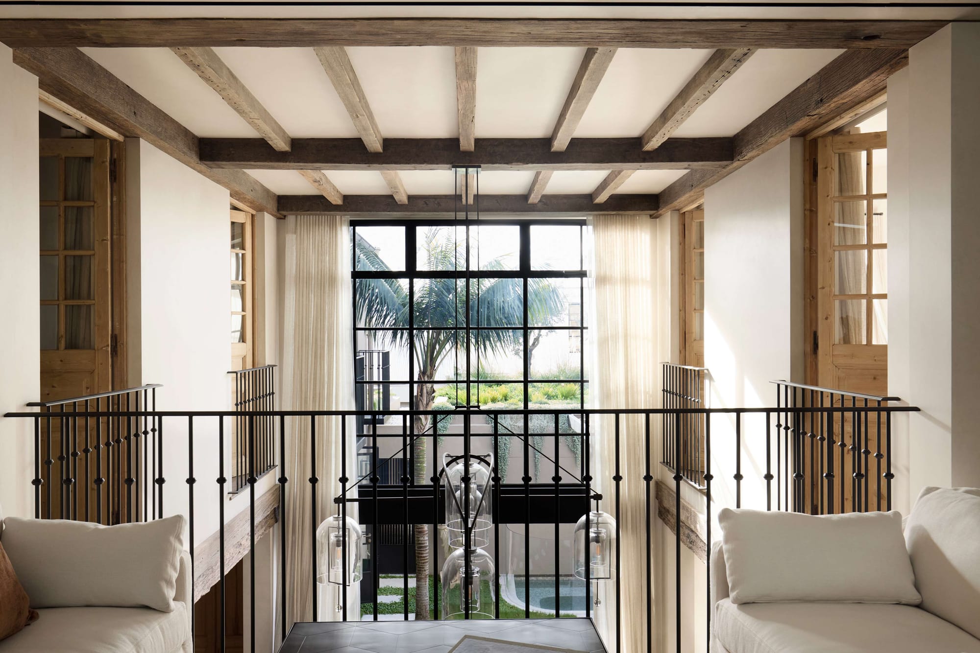 Light-filled interior with exposed timber ceiling beams, black steel balustrades, and double-height glazing overlooking a courtyard garden.